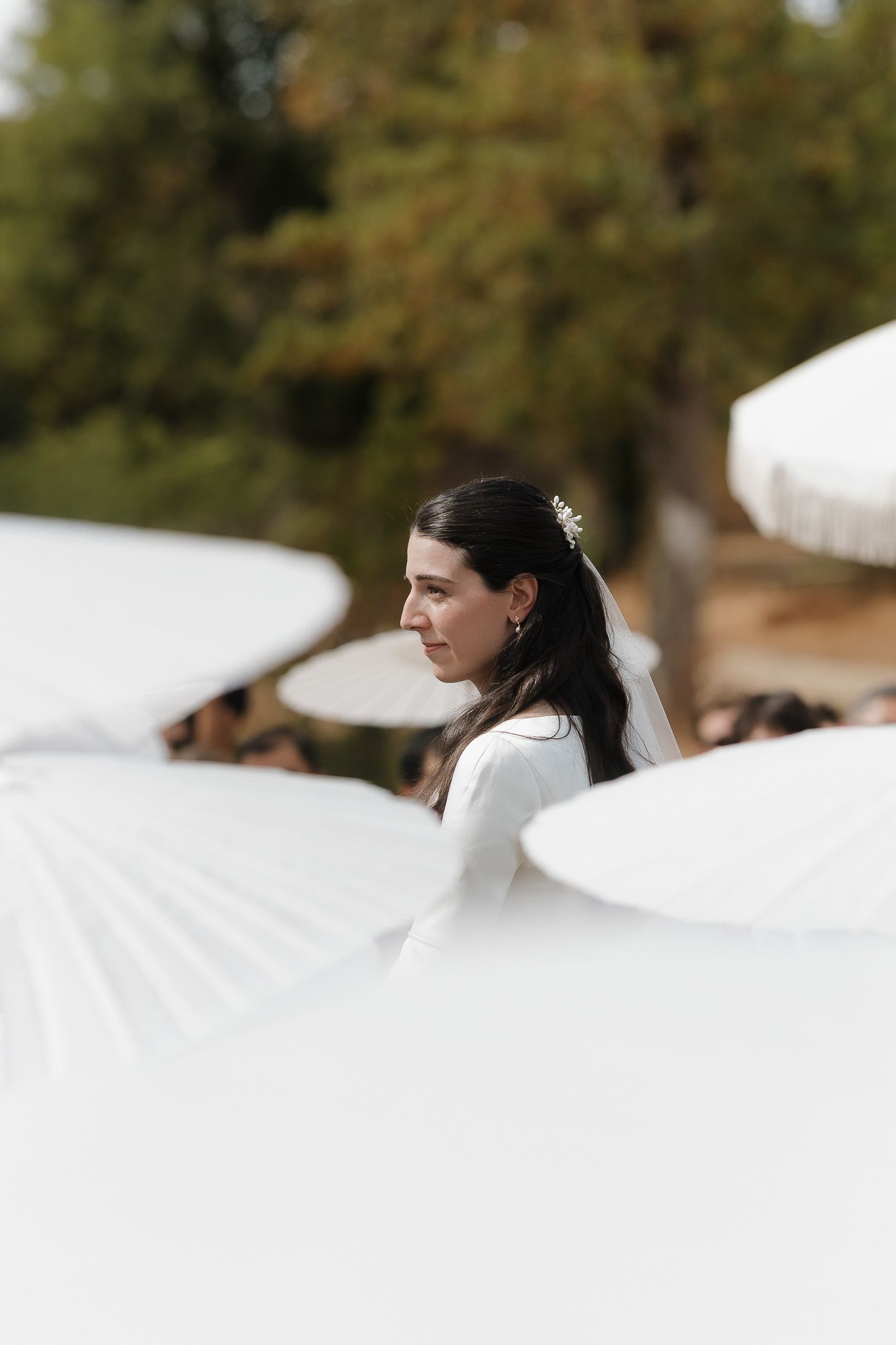 Bride in white dress with floral hair accessory stands among white parasols at an outdoor wedding ceremony.