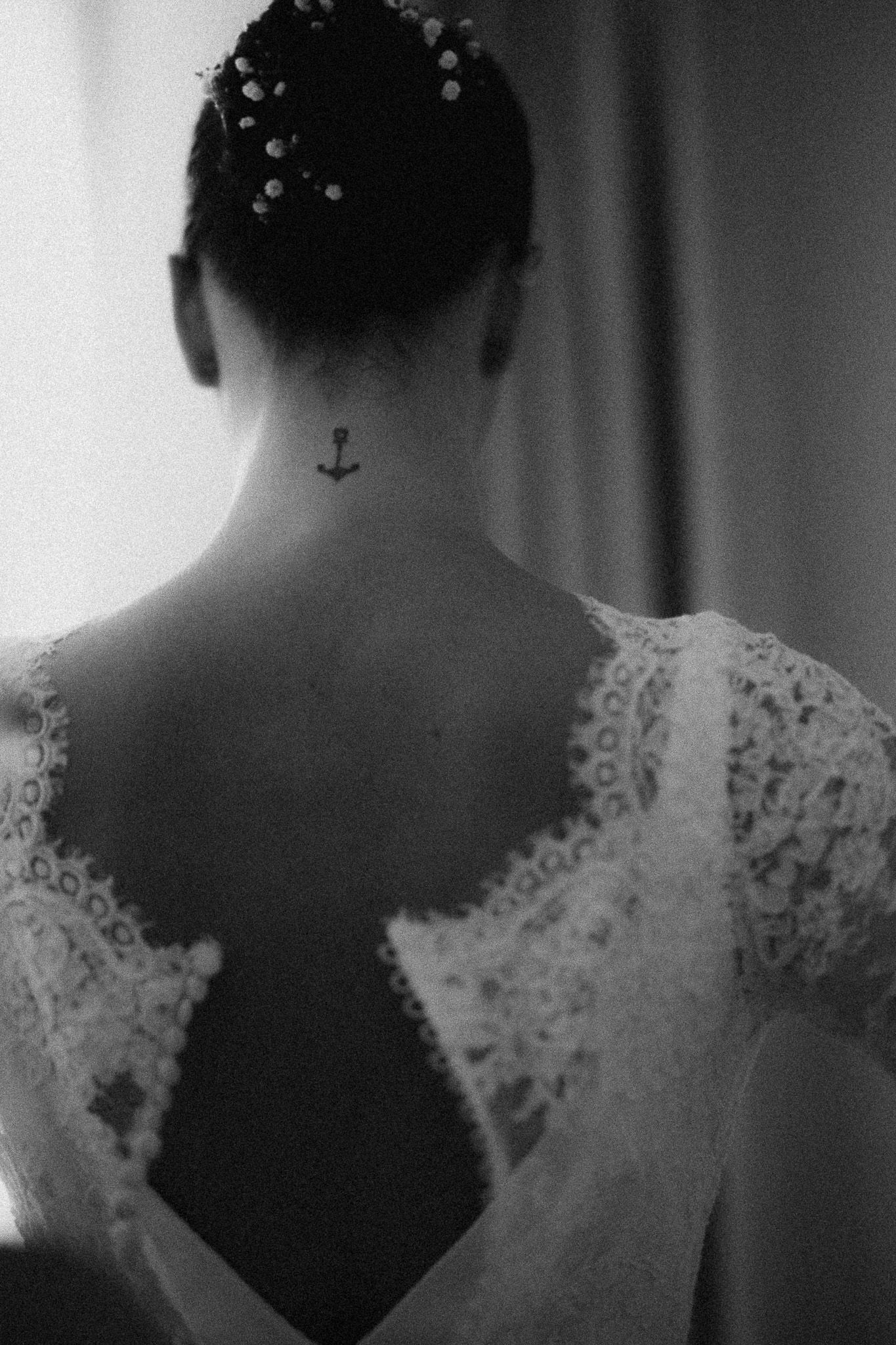 Black and white photo of a bride with an anchor tattoo on her neck, wearing a lace open-back wedding dress.