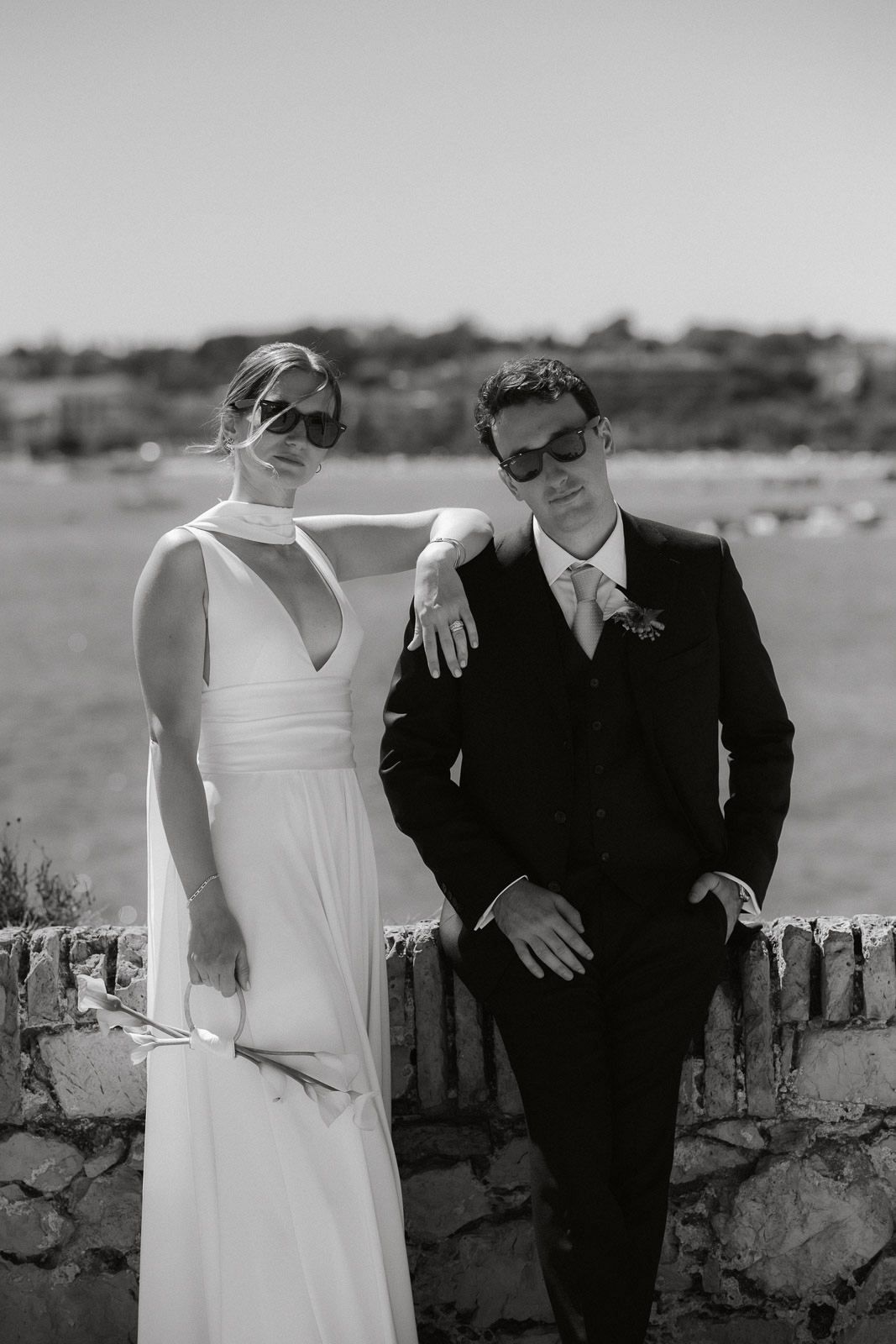 Black and white wedding photo showing couple leaning against stone wall by the ocean.