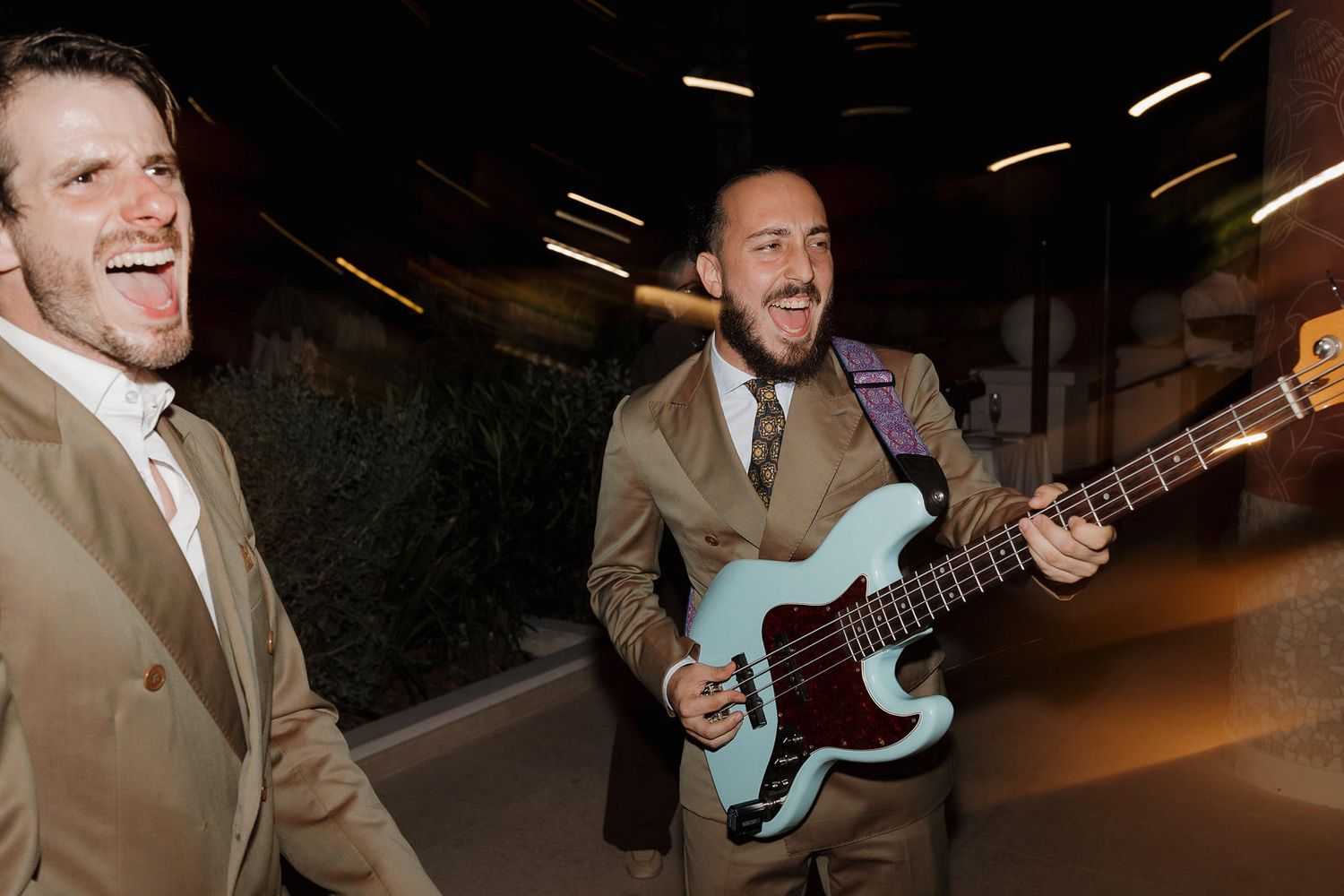Two people in tan suits enjoying a fun moment in a tunnel while one plays a light blue electric bass guitar.