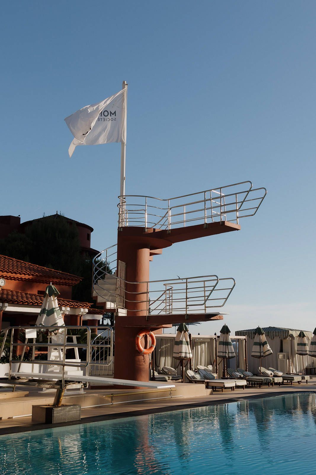 A vintage-style diving platform with white flag overlooks a luxurious hotel swimming pool with lounge chairs.