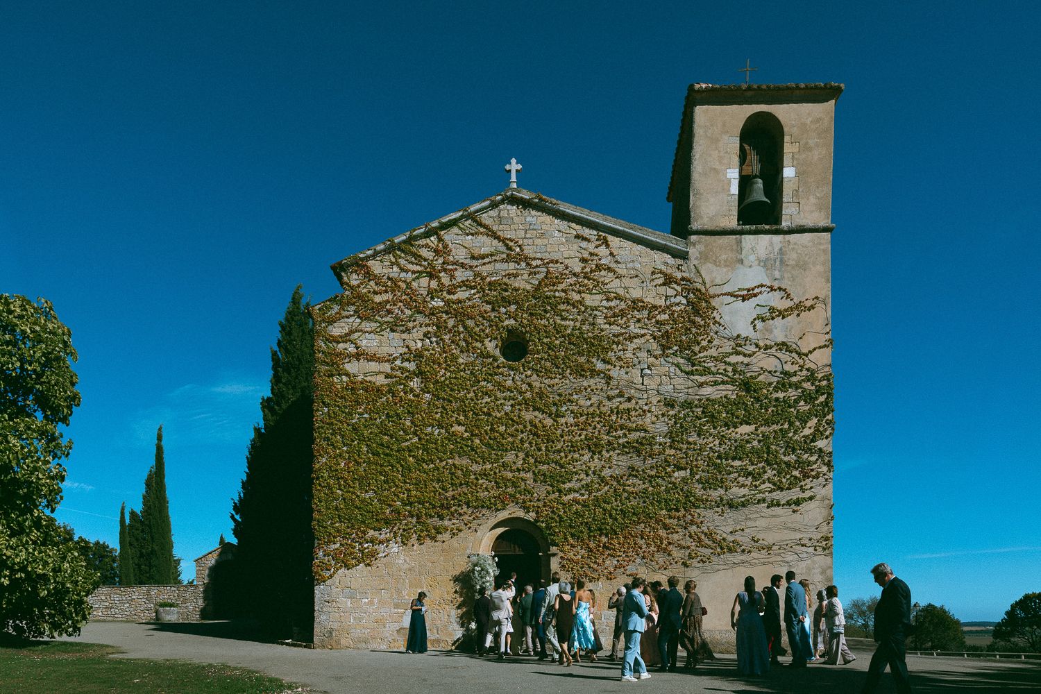 Ivy-covered stone church with bell tower under blue sky, surrounded by cypress trees and visitors.
