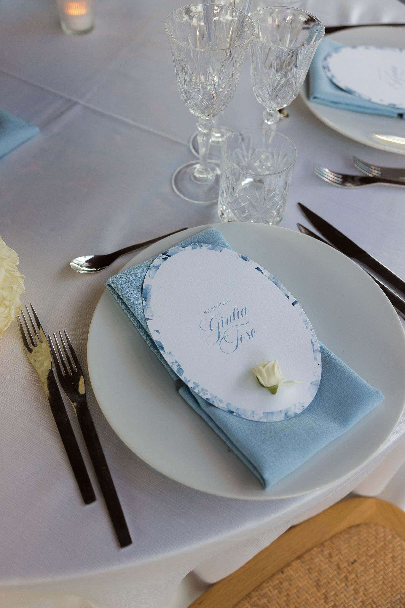 Elegant wedding table setting with blue napkin, place card reading Giulia & Jose, crystal glasses, and white plates.