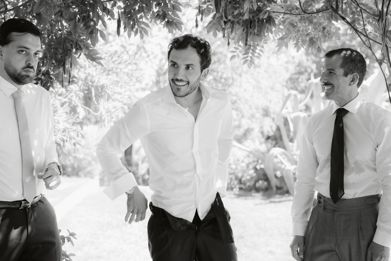 Black and white photo of three men in dress shirts smiling and laughing together outdoors at a wedding.