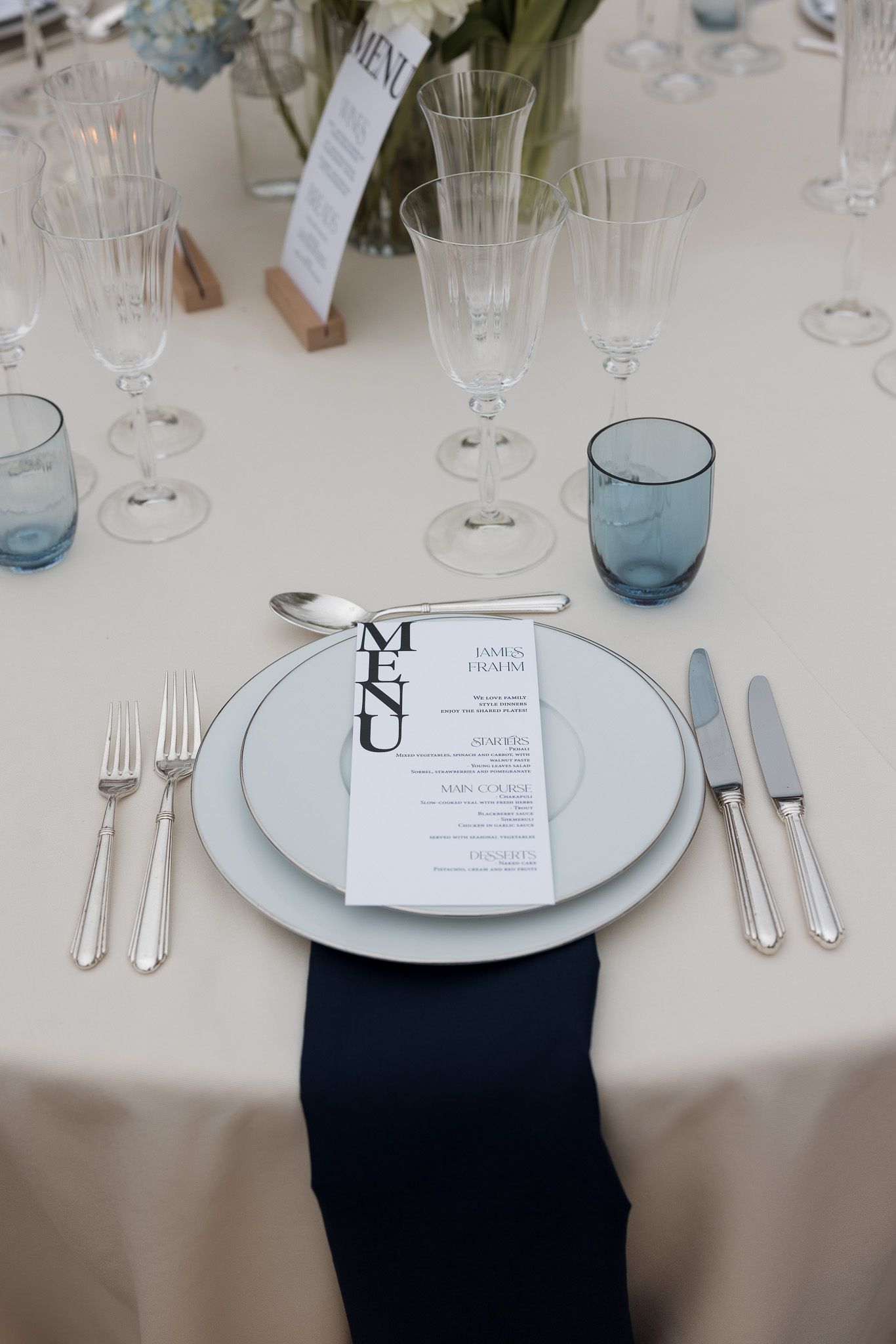 Elegant wedding table setting with navy napkin, crystal glasses, silver cutlery, and a menu card on white plates.