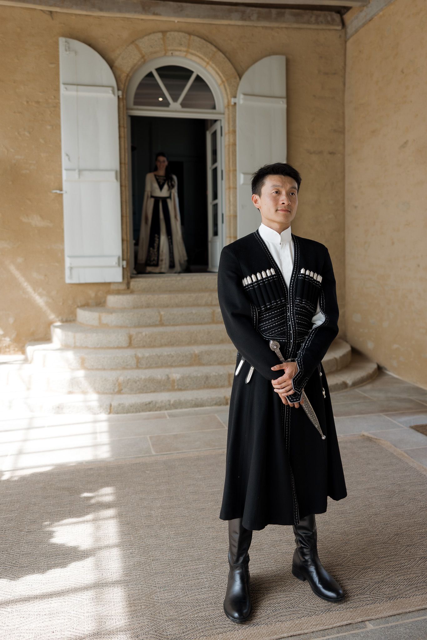 Man in traditional black Georgian chokha costume holding a dagger stands before a historic building entrance.