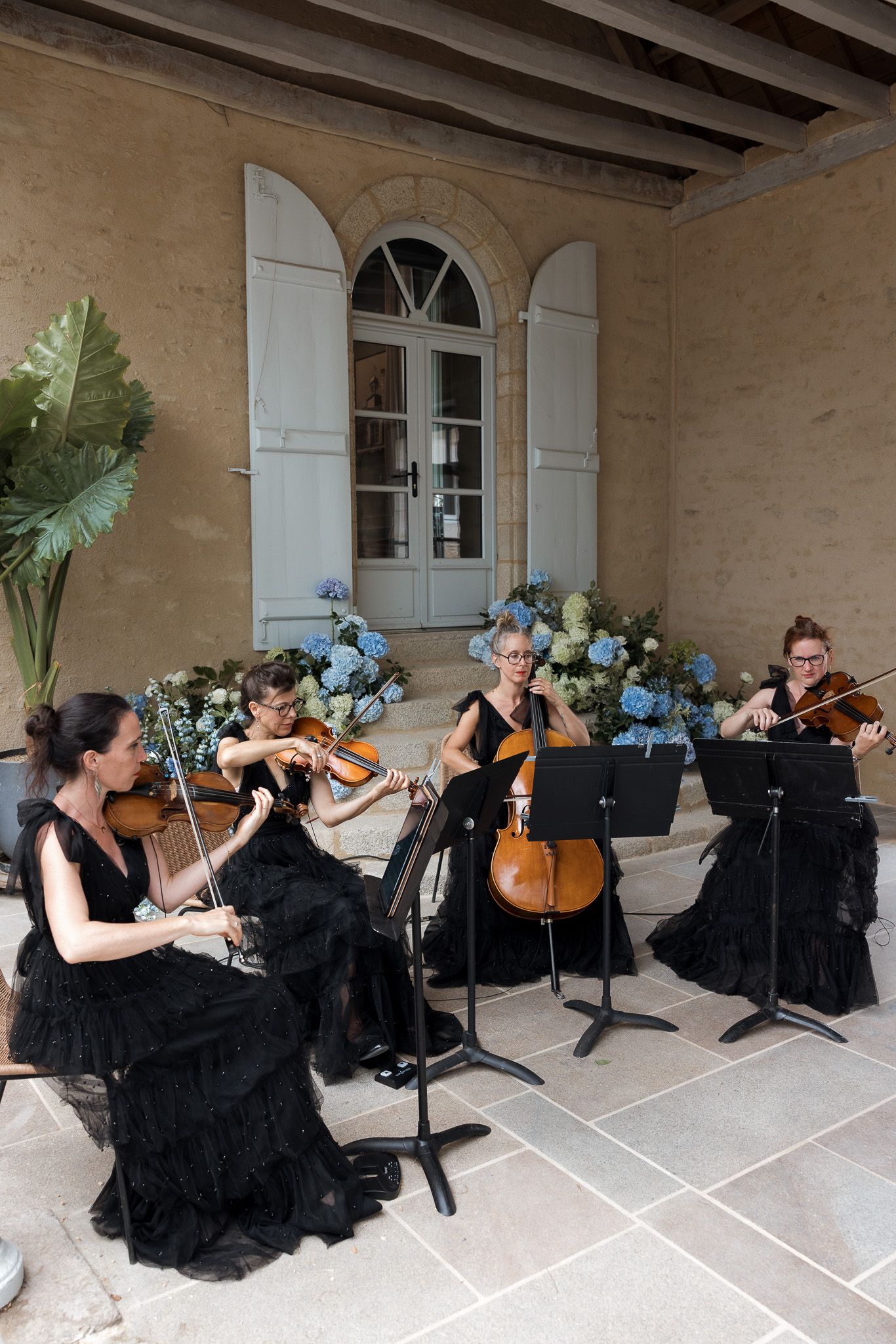 String quartet in black gowns performing at an elegant outdoor venue with floral arrangements and stone architecture.