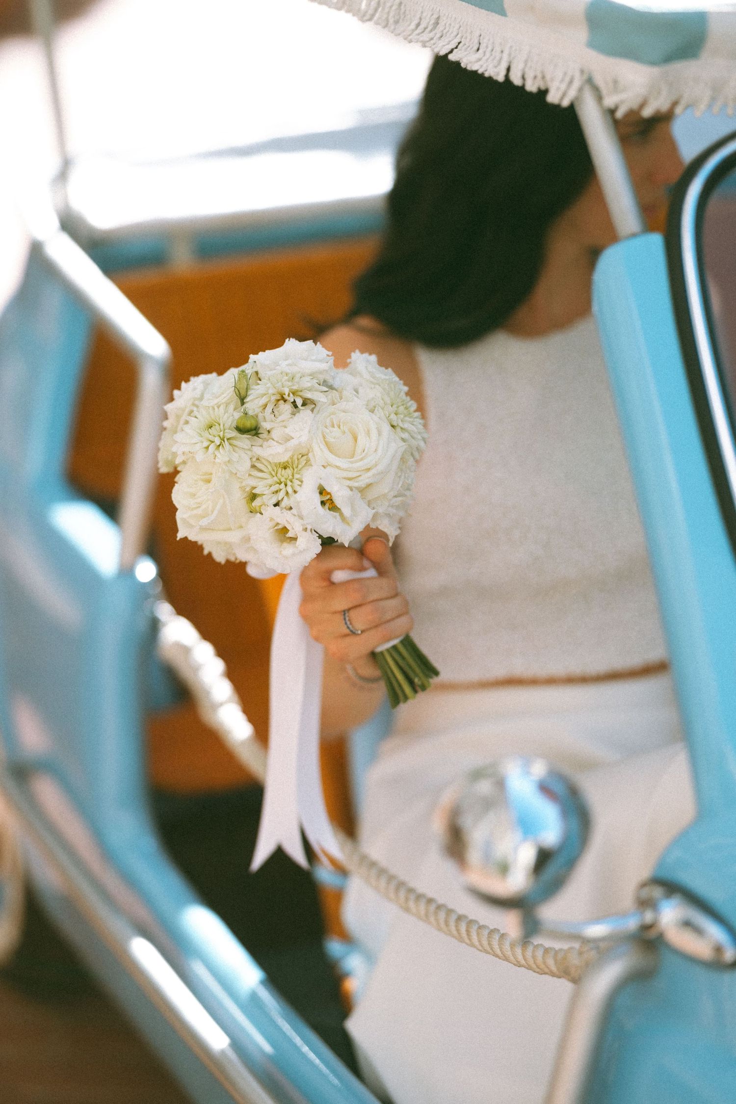 Bride holding a white rose bouquet with ribbon while seated in a vintage turquoise car on her wedding day.