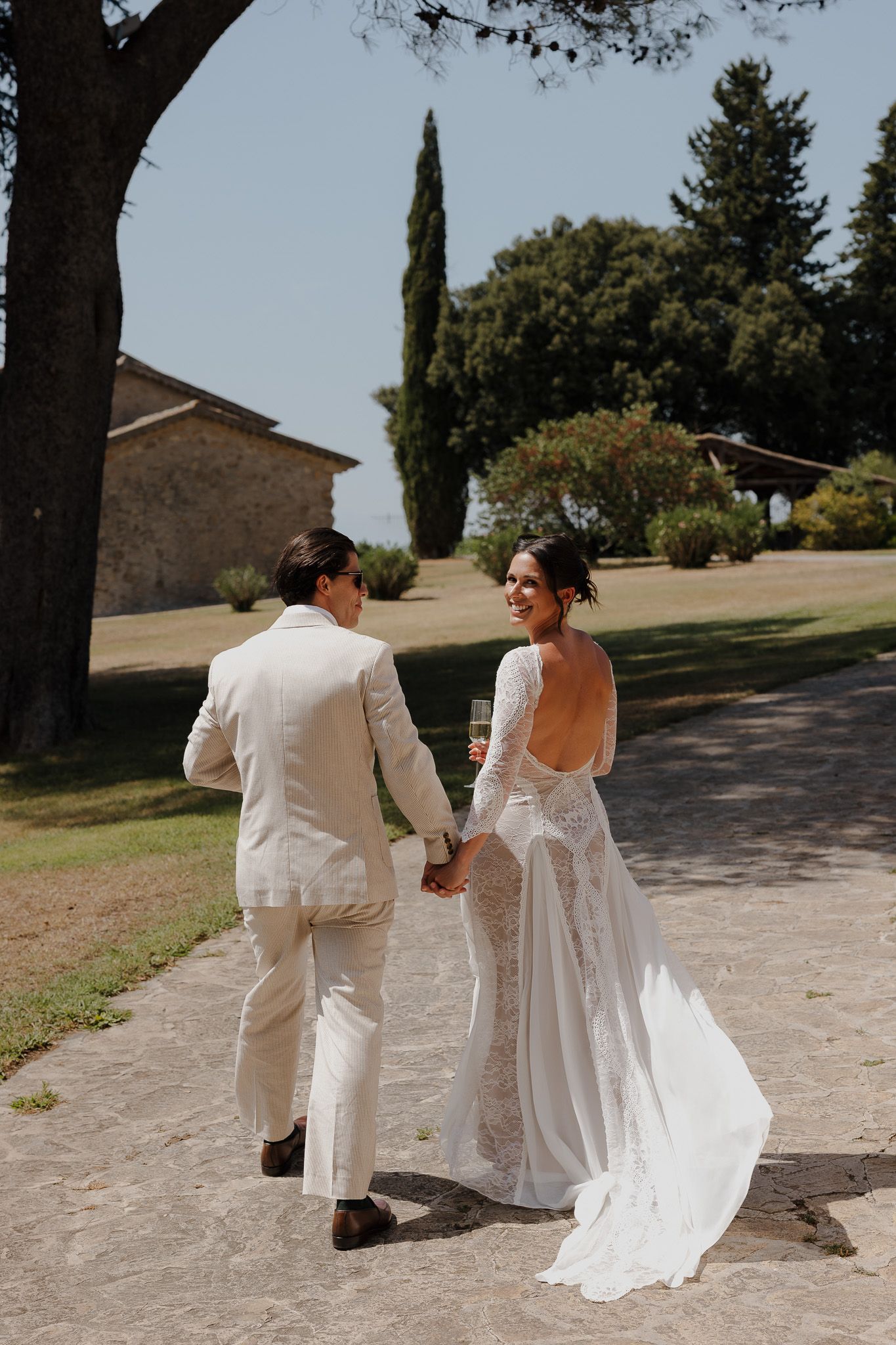 Bride in backless lace gown holds hands with groom in cream suit, walking through Tuscan villa grounds.