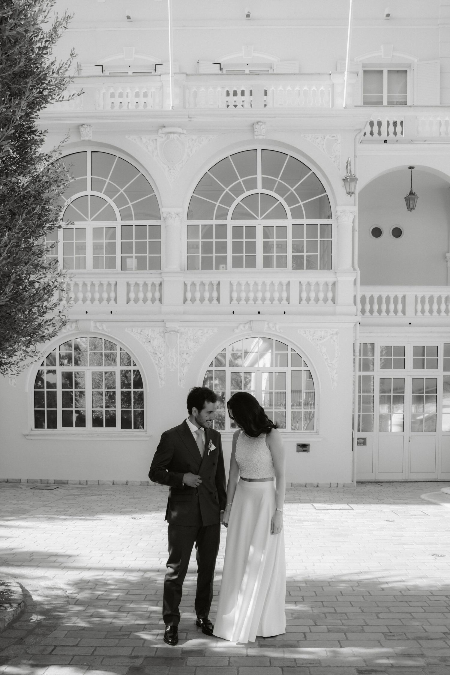 Black and white photo of a couple in wedding attire standing before an elegant white building with arched windows.