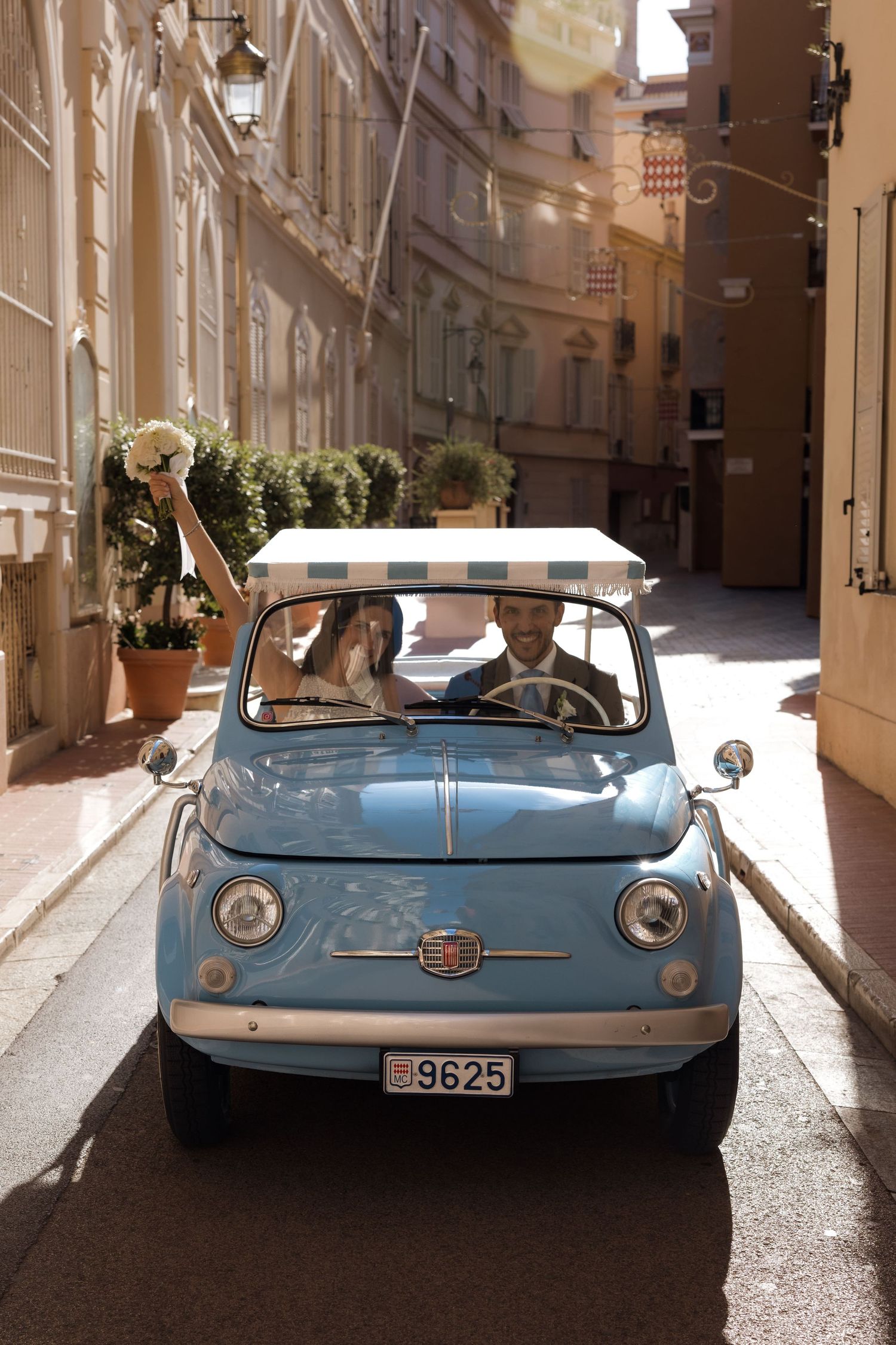 Newlyweds celebrate in a vintage baby blue Fiat 500 driving through sunny European streets, bride holding bouquet aloft.