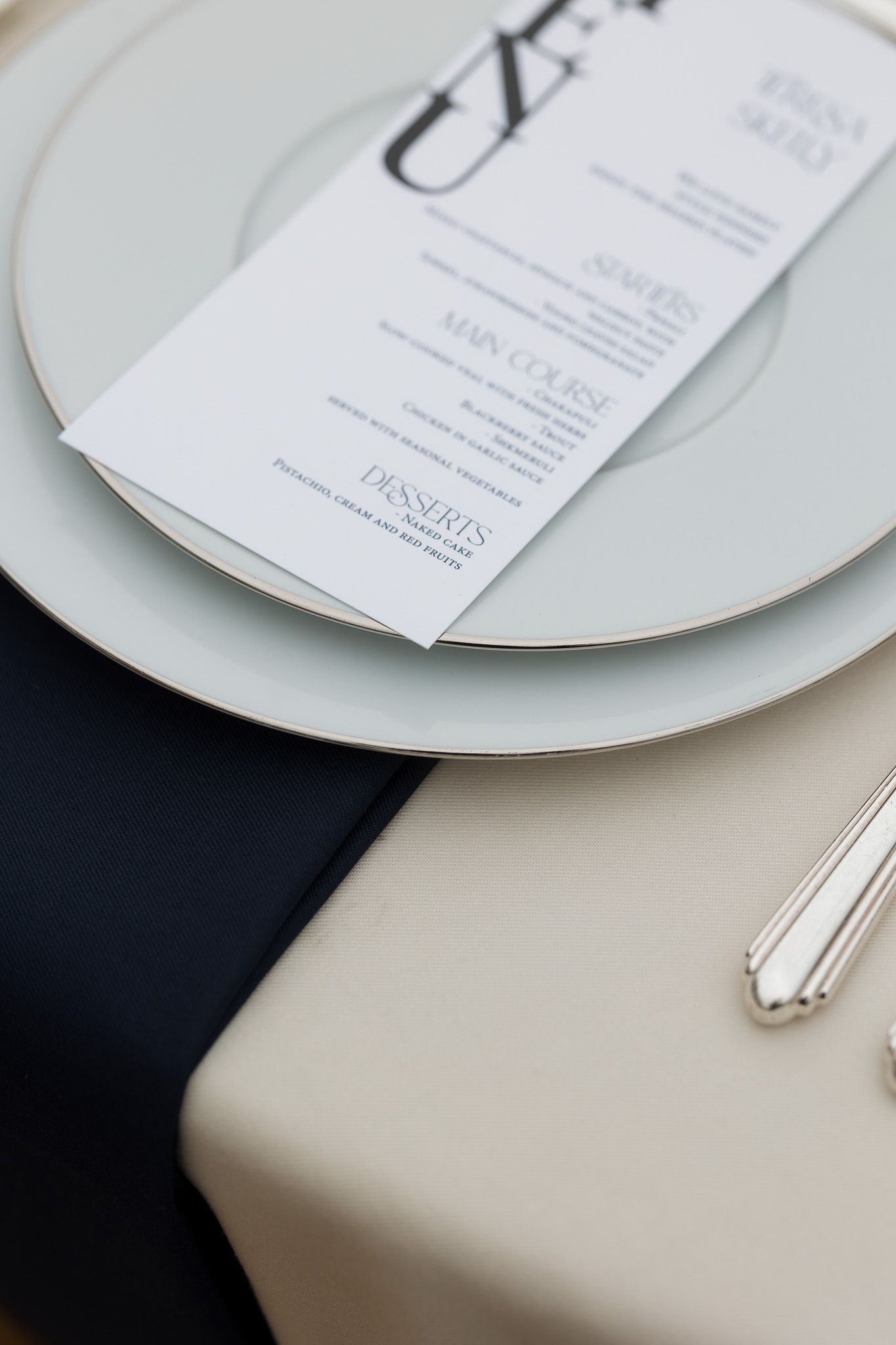 Elegant wedding place setting with white plates, silver cutlery, black napkin, and a printed menu card on a cream tablecloth.