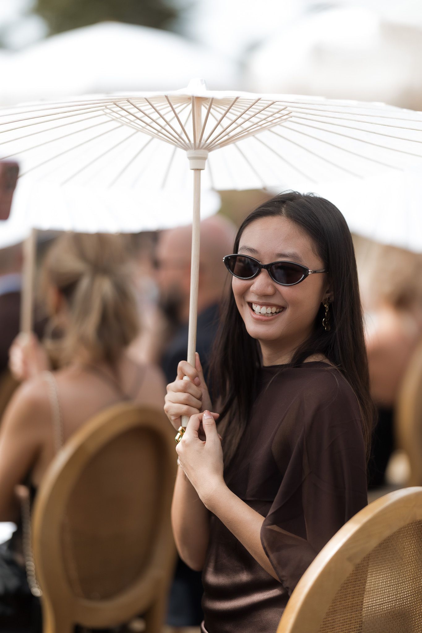 Smiling woman in sunglasses holding a white parasol umbrella at an outdoor social event.