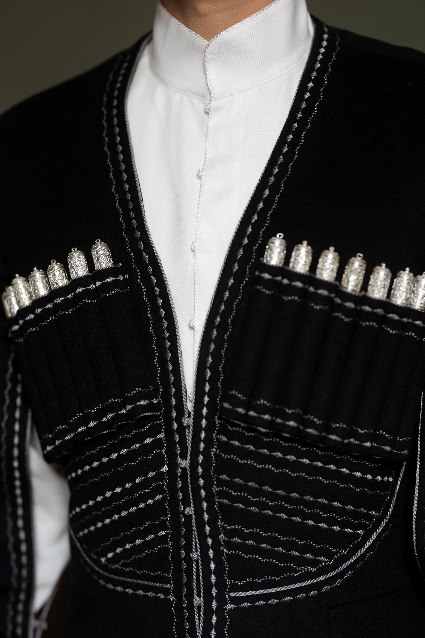 Black Circassian Chokha coat with silver gazyr cartridge holders and white embroidery detailing over white shirt.