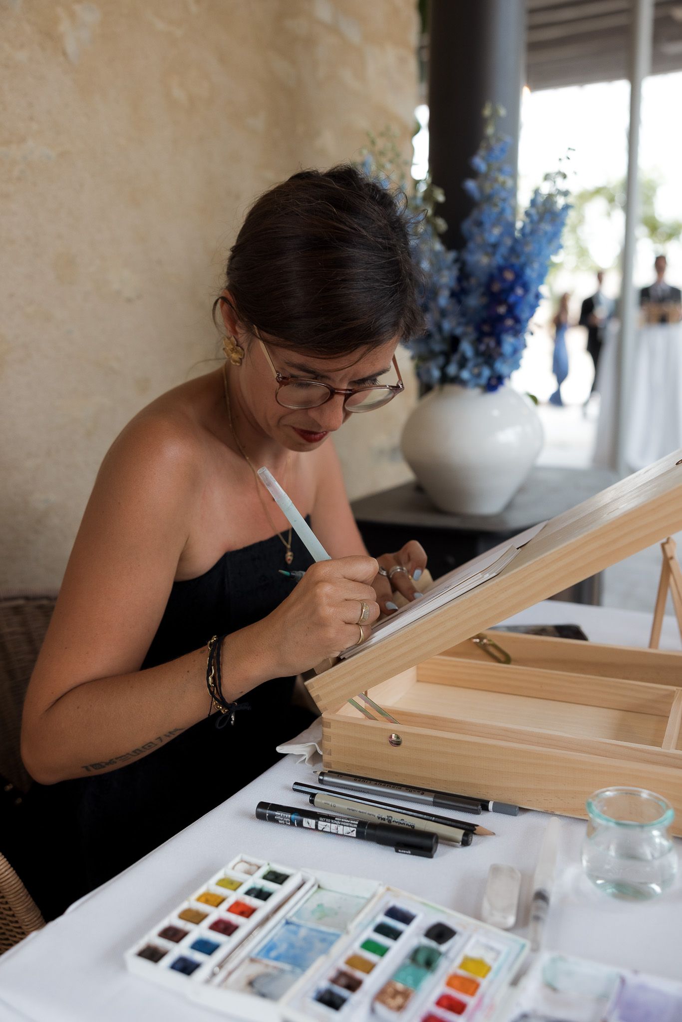 Woman in black dress painting with watercolors at an easel, wearing glasses, with art supplies on table.