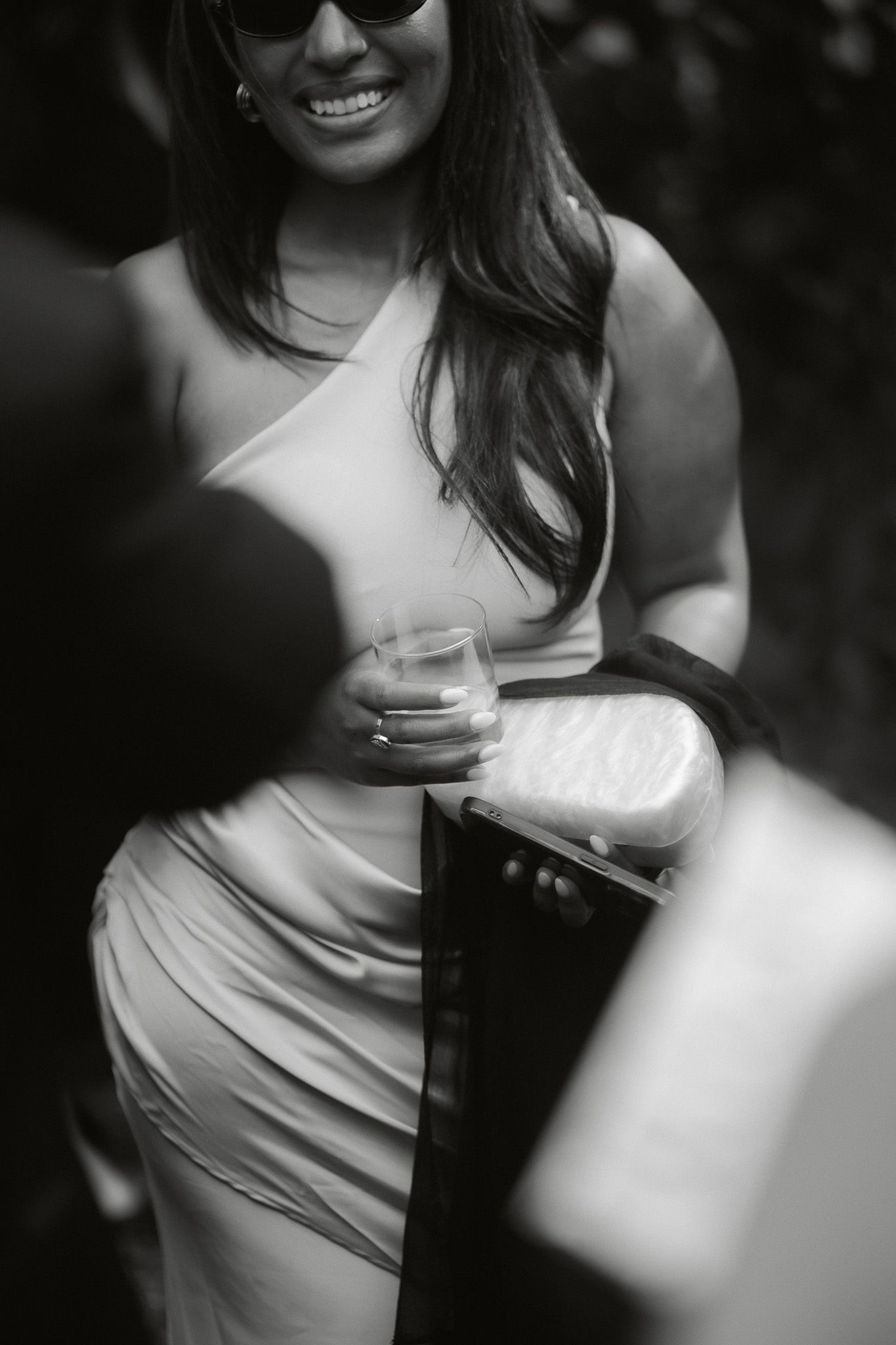 Smiling woman in elegant dress holding a glass at an outdoor event, black and white photography.