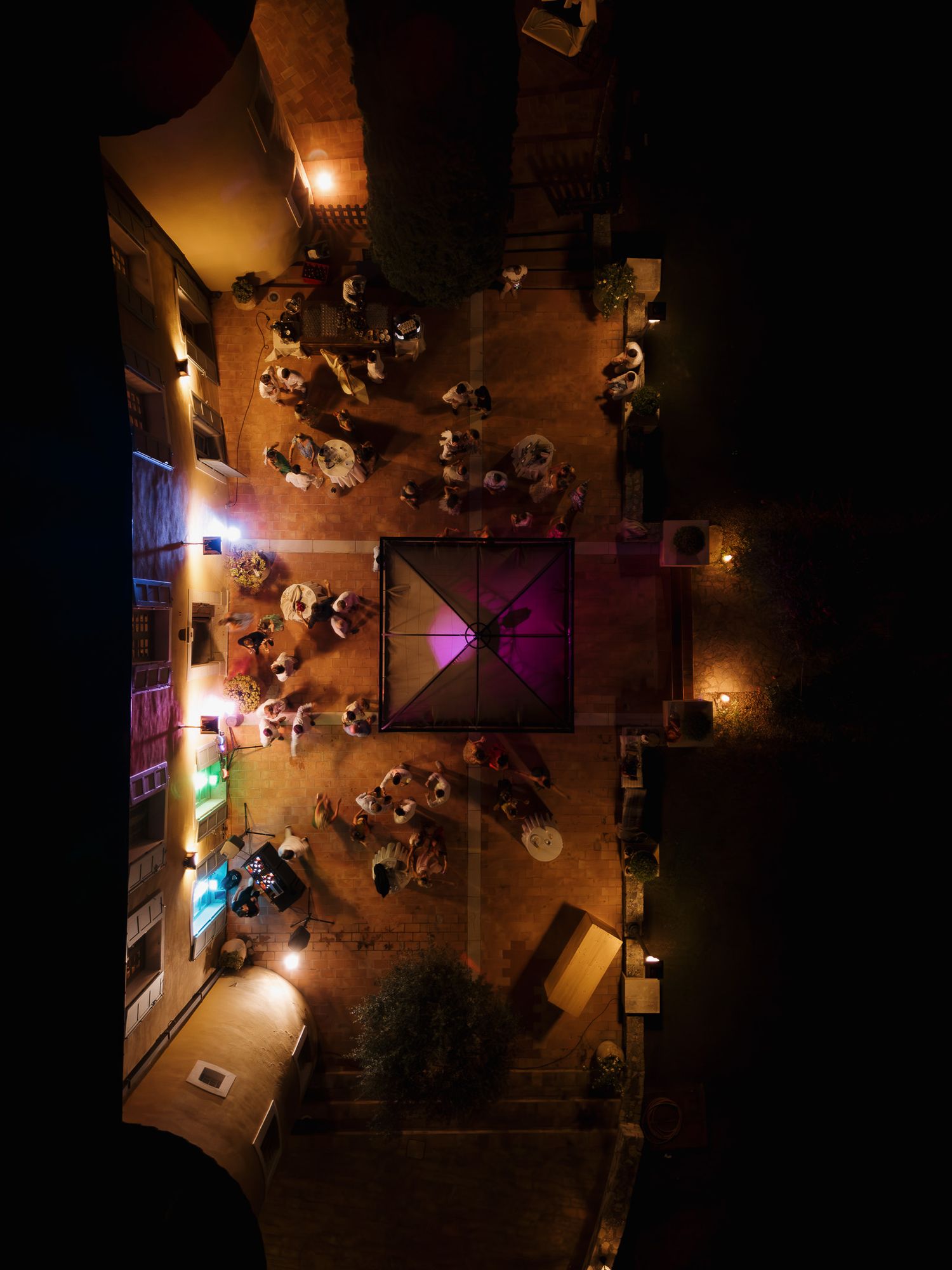 Aerial night view of an outdoor courtyard event with warm lighting, glowing purple centerpiece, and guests mingling around tables.