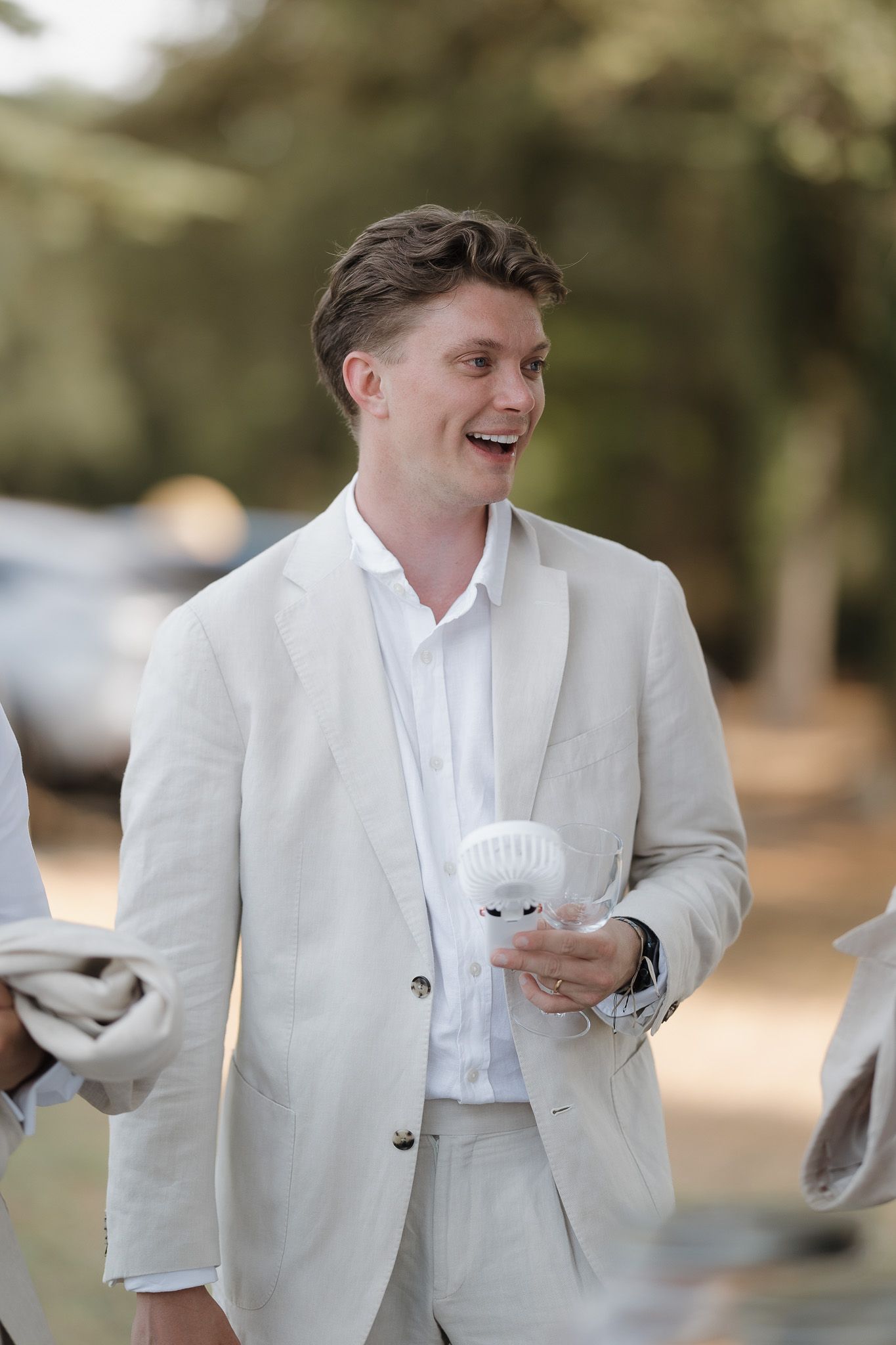 Smiling young man in a white linen suit holding a cup outdoors at a garden event.