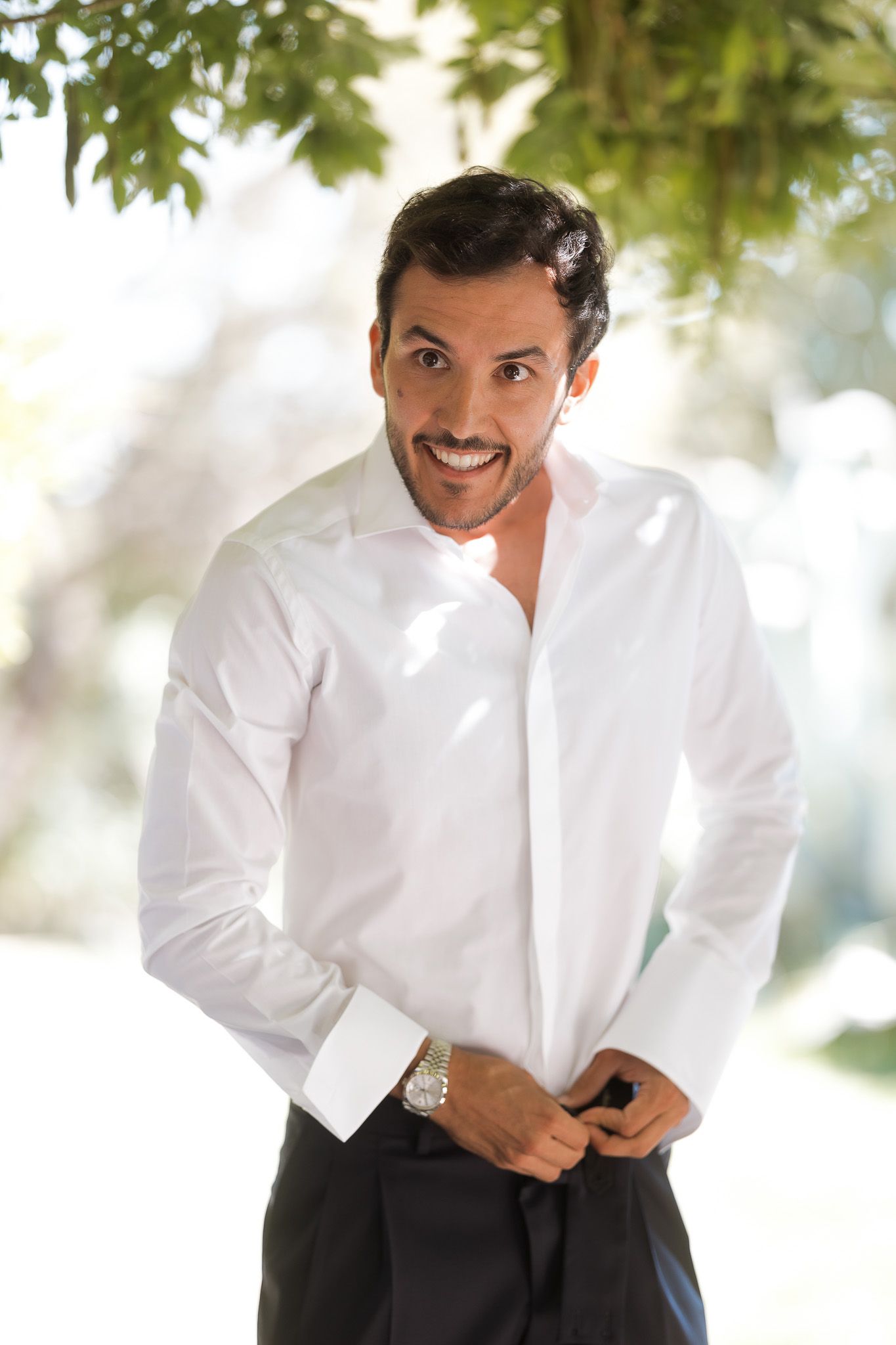 Smiling man in white dress shirt and black pants adjusting his clothing outdoors near green trees.