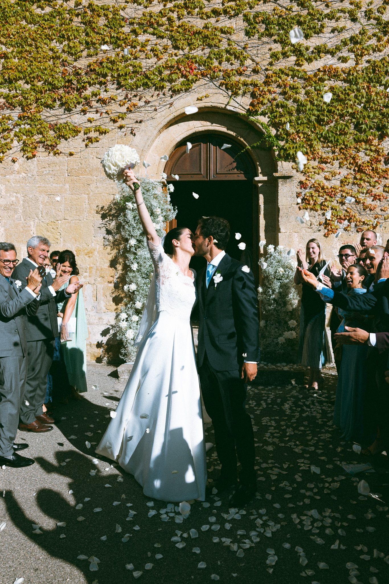 Bride raises bouquet while groom kisses her cheek outside stone church, surrounded by guests tossing white flower petals.