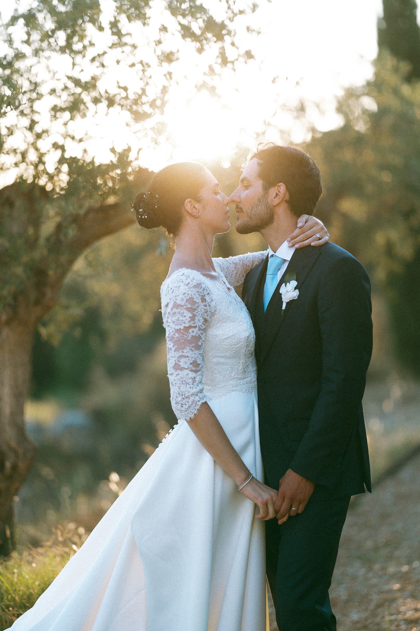 Bride and groom share a romantic kiss at golden hour, surrounded by olive trees, as sunlight glows behind them.