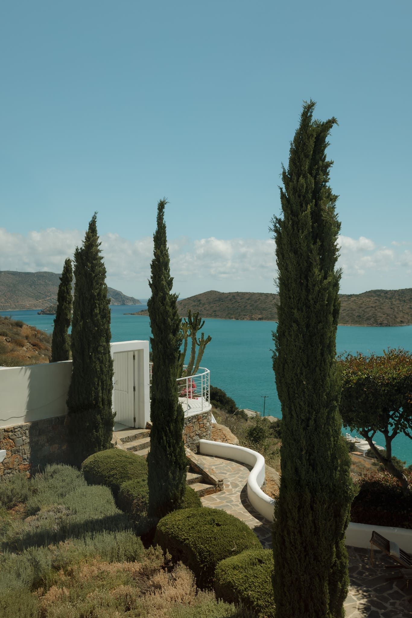 Mediterranean coastal landscape with tall cypress trees and white buildings overlooking turquoise waters and distant hills.