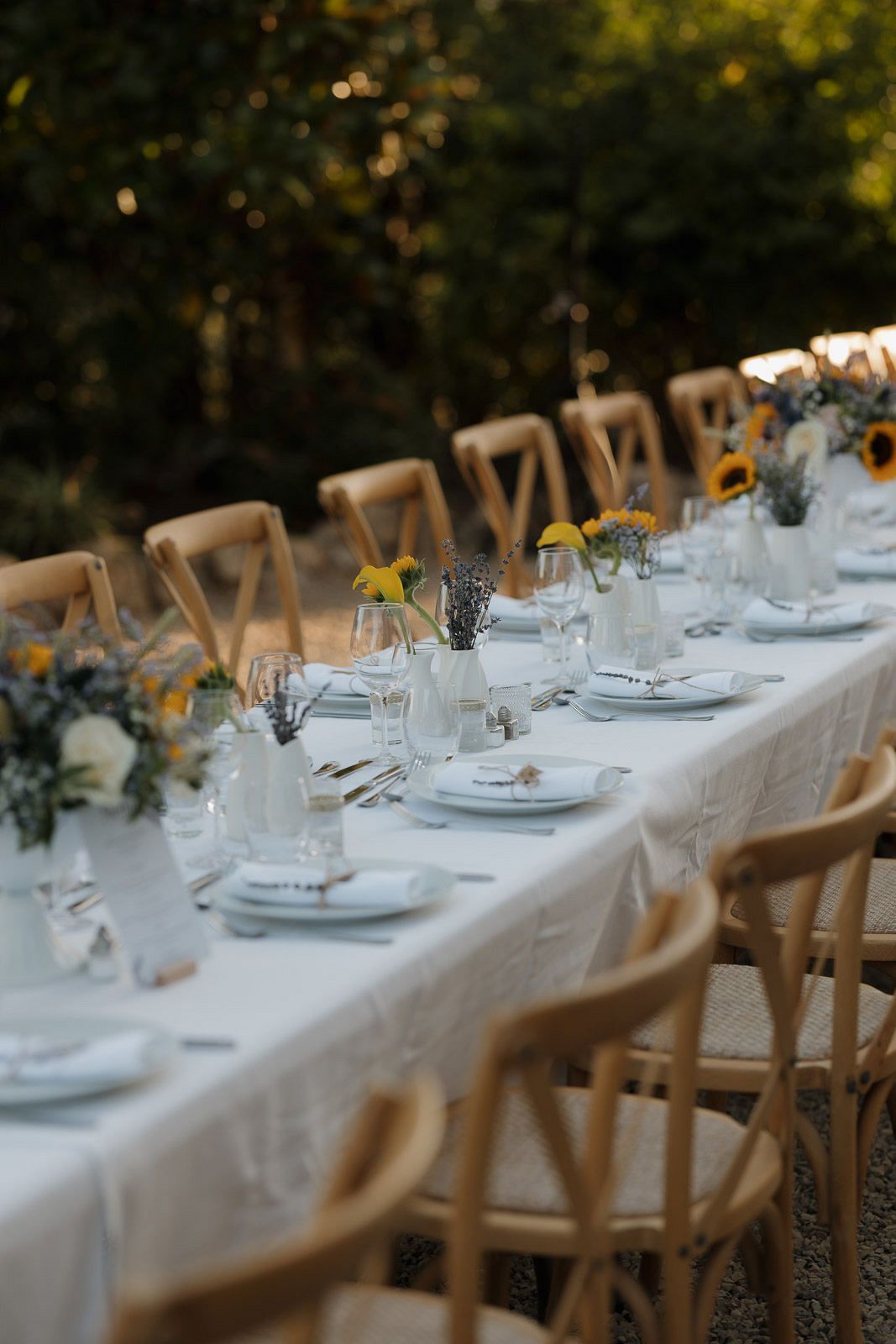Long white dining table set with elegant place settings and wooden chairs for an outdoor garden party.