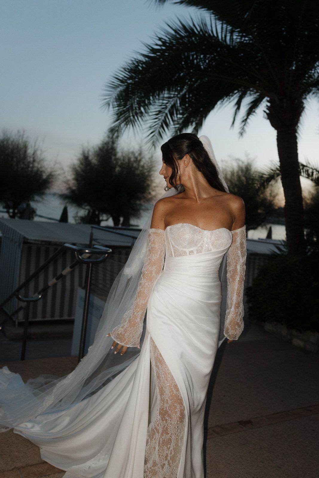 A bride in a white wedding dress with lace sleeves poses against a backdrop of palm trees at dusk.