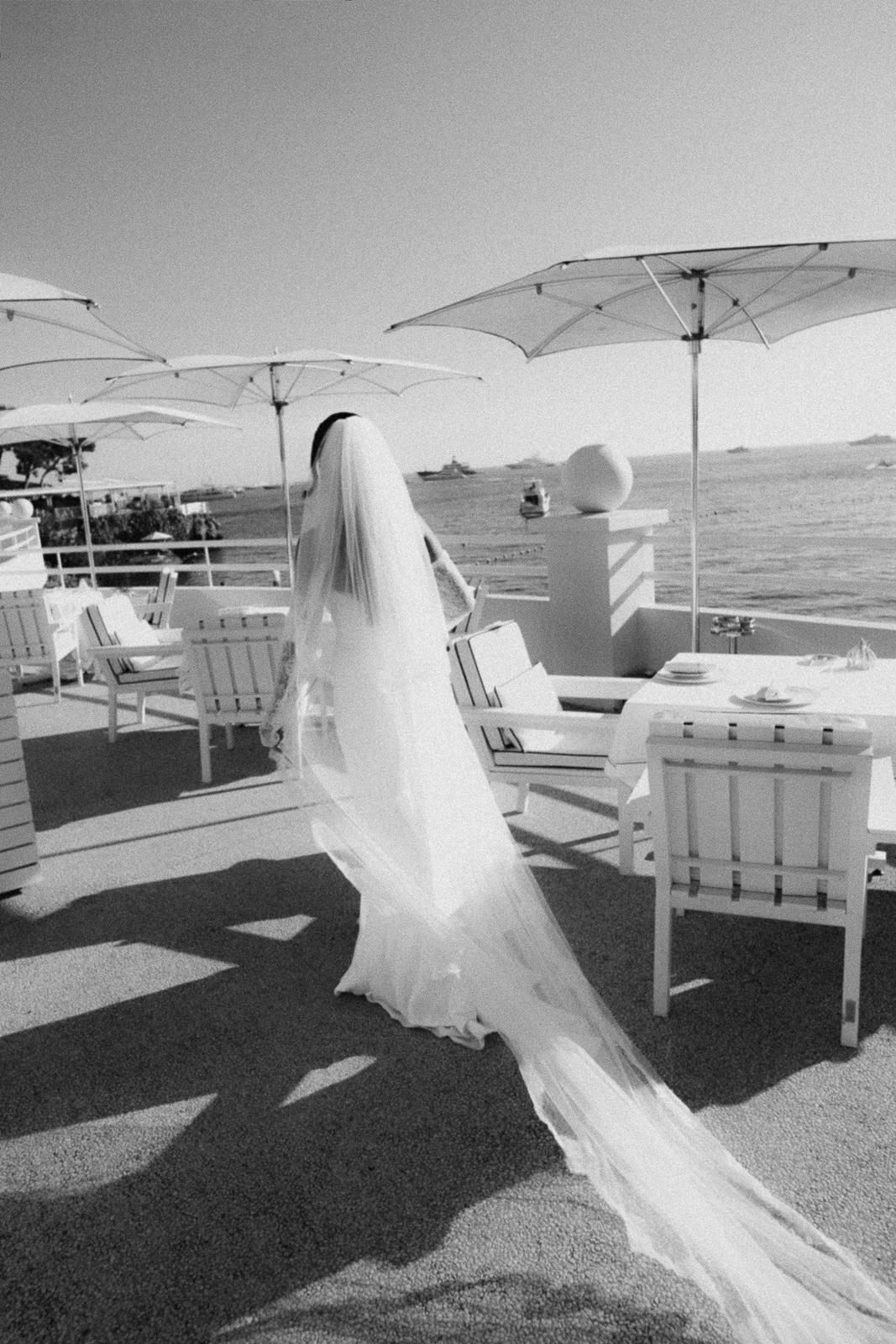A flowing white veil billows in the wind across white outdoor furniture and umbrellas at a scenic outdoor venue.