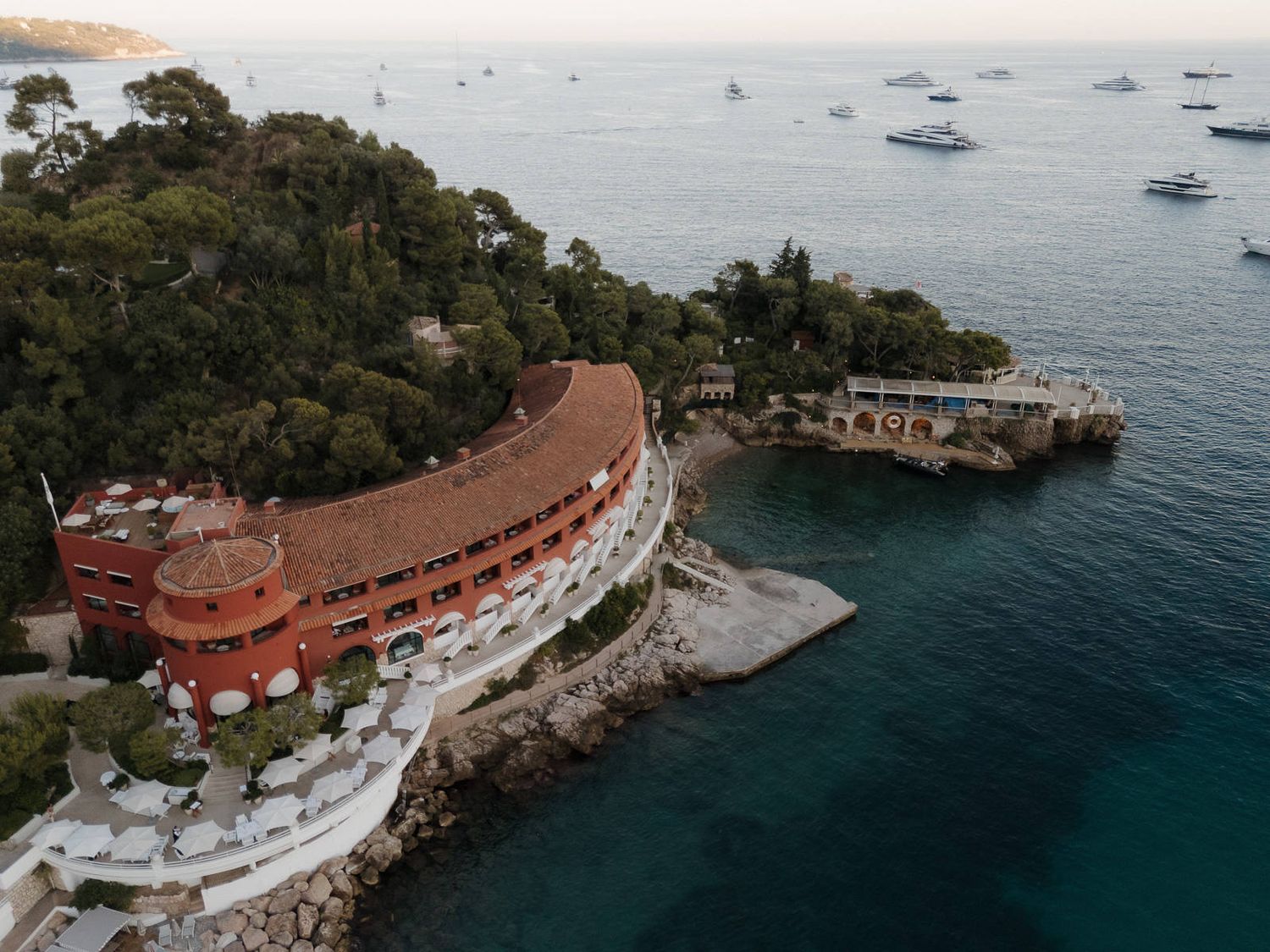Aerial view of a curved luxury hotel perched on a cliff overlooking the Mediterranean Sea with yachts anchored in the bay.
