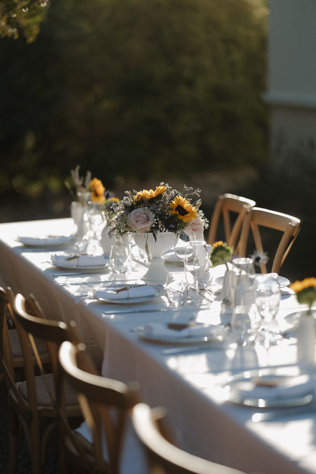 A long white dining table set with elegant place settings and sunflower centerpieces in natural outdoor lighting.