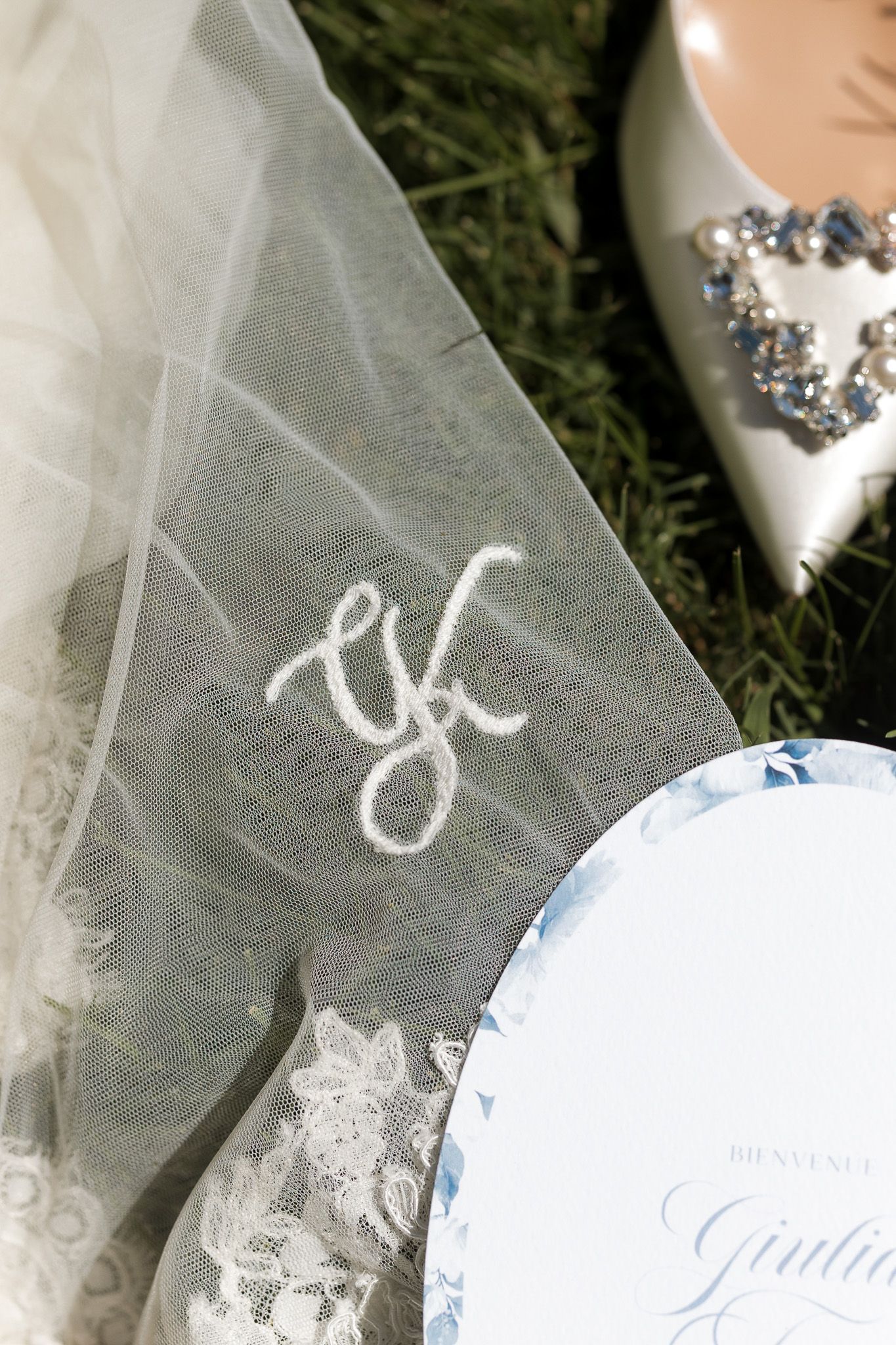 Monogrammed wedding veil with lace trim beside a blue floral invitation and jeweled bridal shoe on grass.