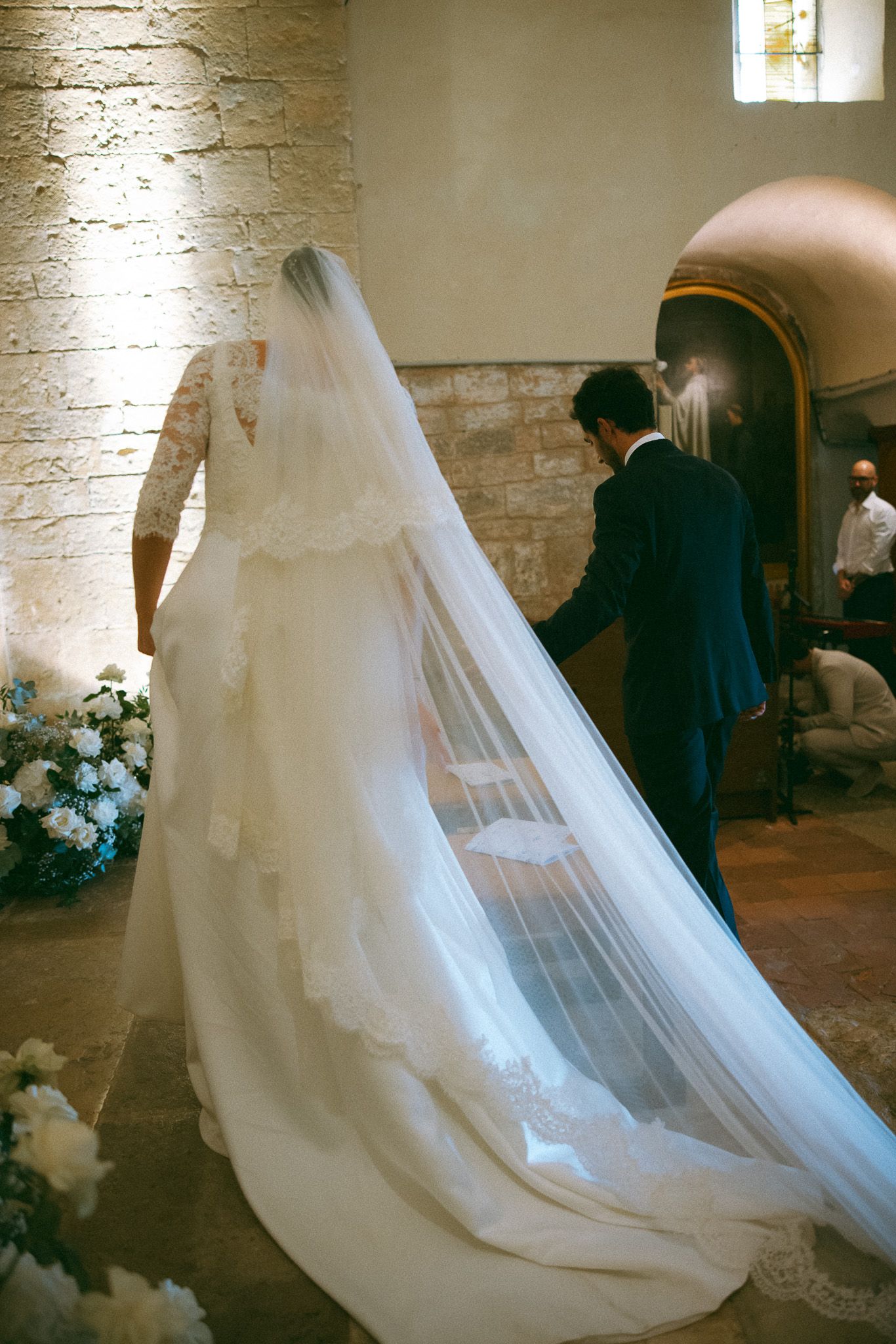 Bride in lace wedding gown with long cathedral veil stands beside groom in blue suit inside stone church.