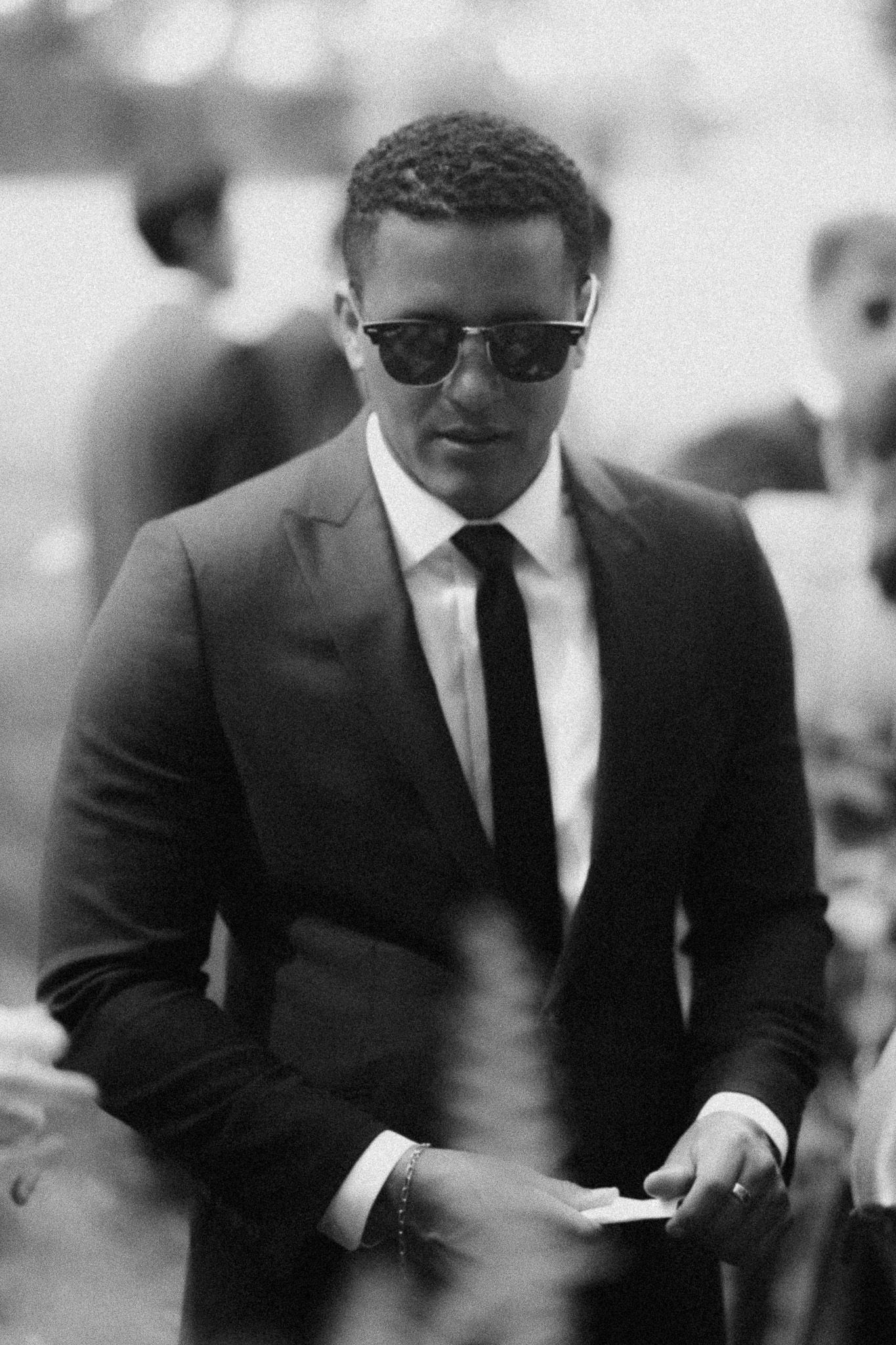 Black and white photo of a stylish man in a suit and tie wearing sunglasses at an outdoor event.