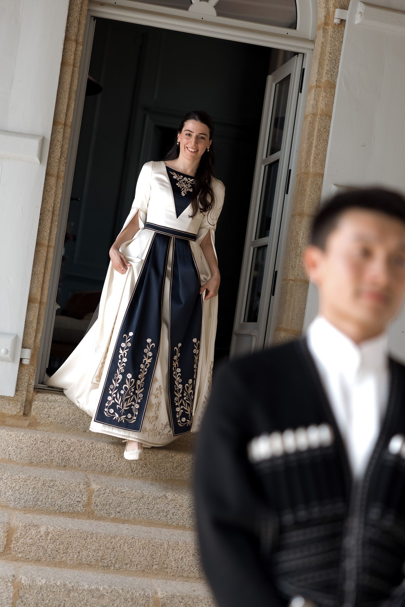 Woman in elegant white and navy embroidered gown descends steps of grand building as man in suit stands in foreground.