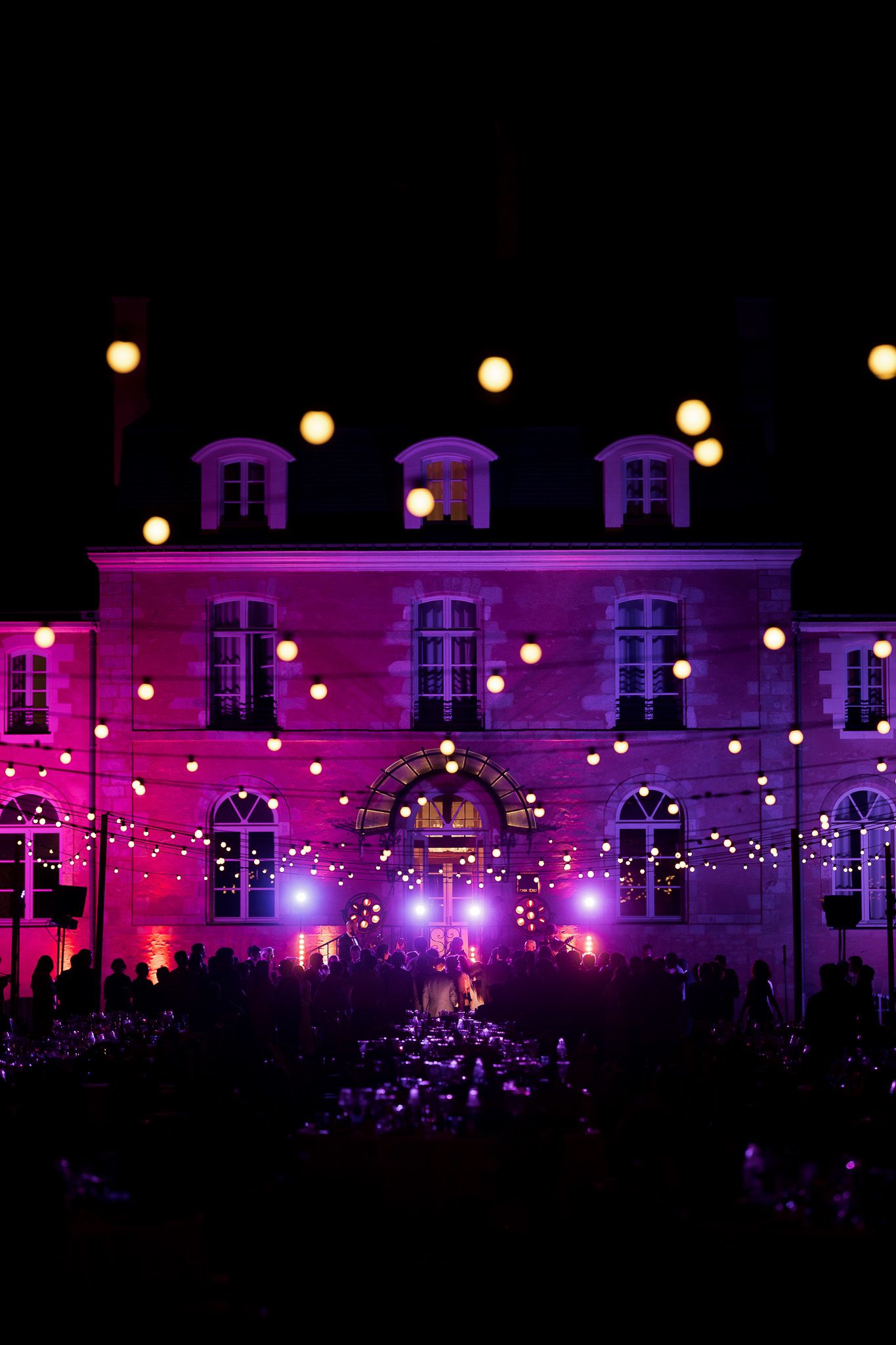 A stunning French chateau illuminated in purple light with string lights during an elegant outdoor wedding reception at night.