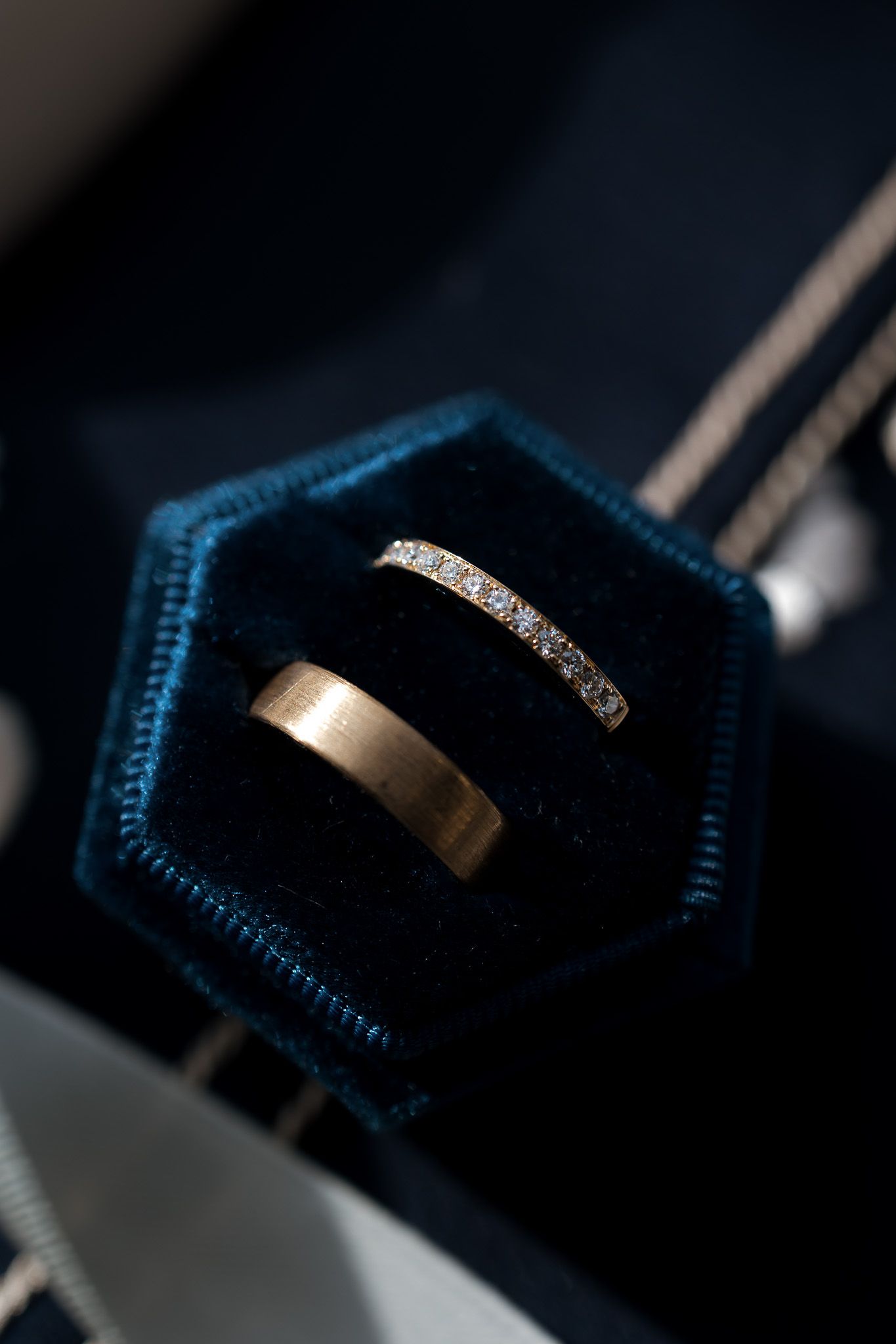 Two gold wedding rings, one diamond eternity band, displayed in a teal velvet hexagonal ring box.