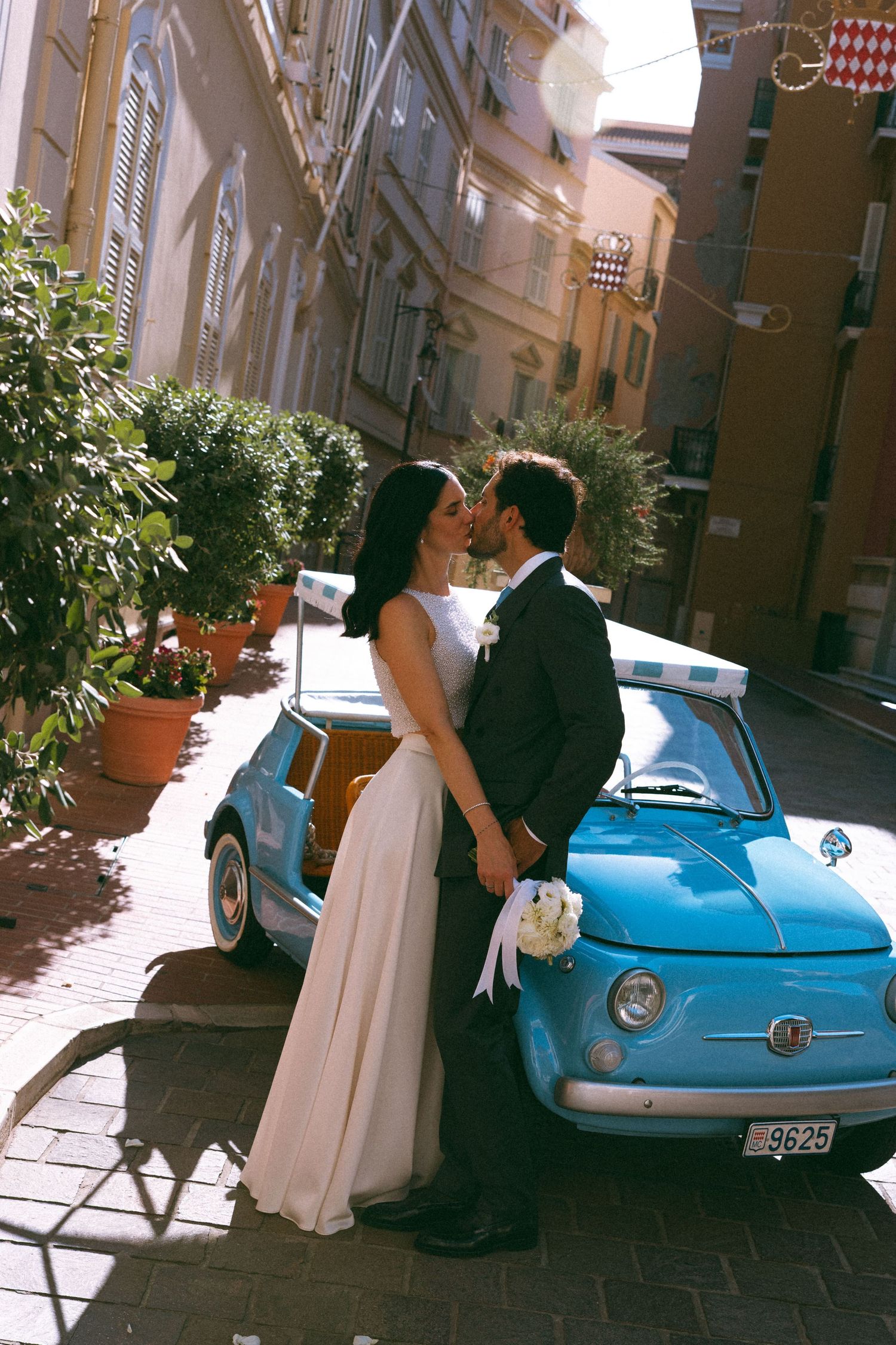 Newlyweds kissing beside a vintage blue Fiat 500 on a sunlit European cobblestone street, bride holding white bouquet.