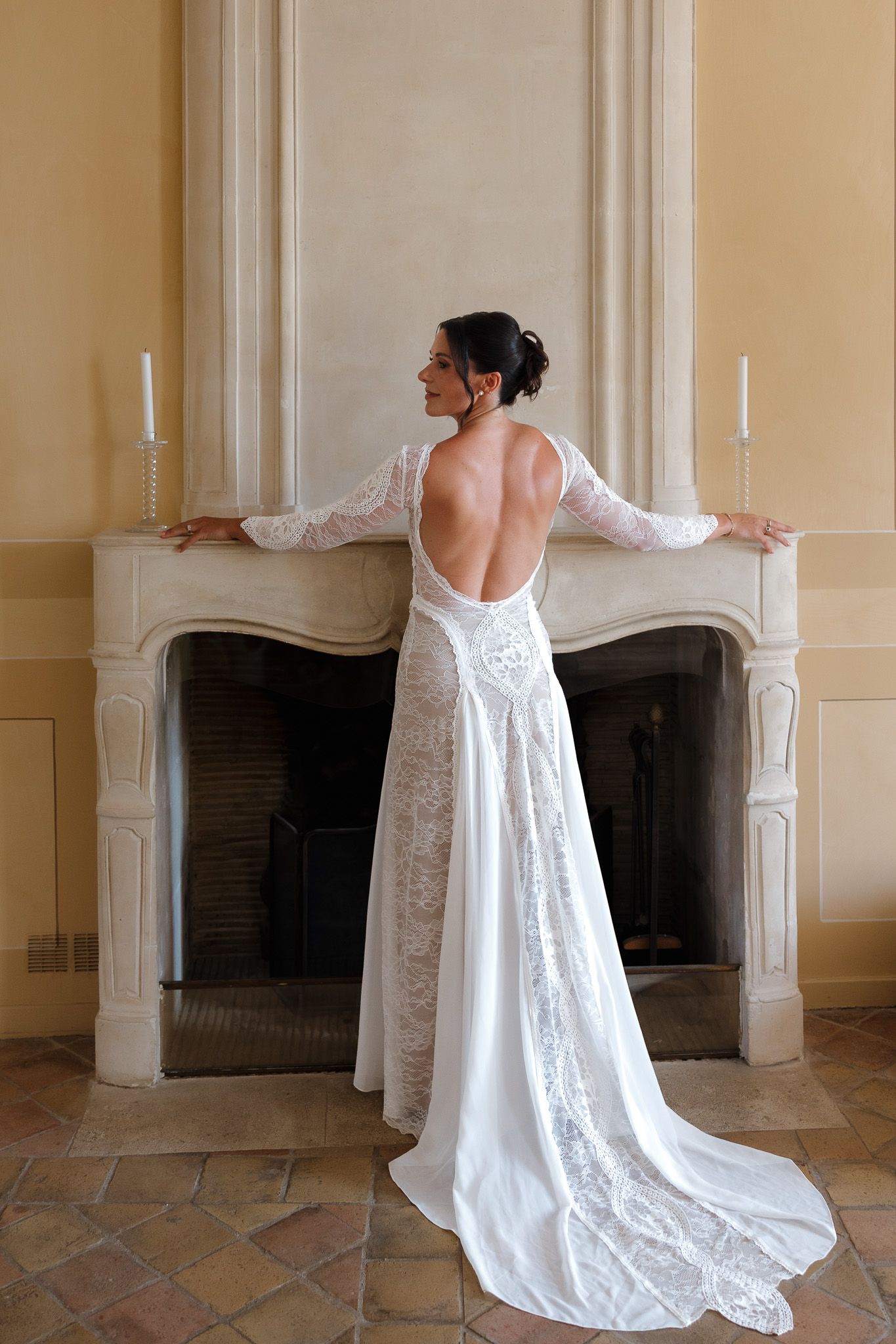 Bride in elegant backless lace wedding gown with train stands at ornate fireplace with candles.