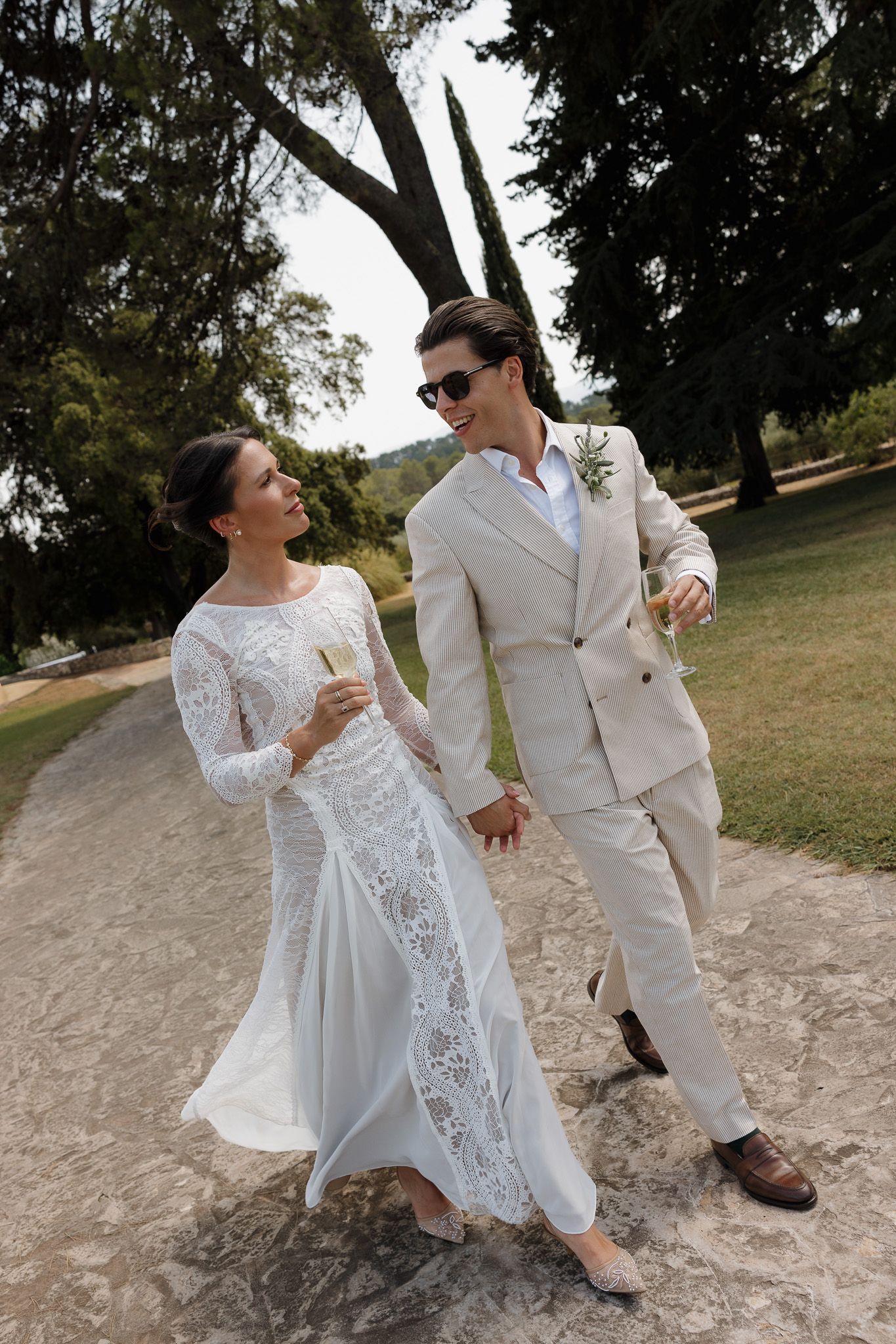 Bride and groom walking hand in hand outdoors, she in lace gown, he in beige suit, both holding champagne glasses.