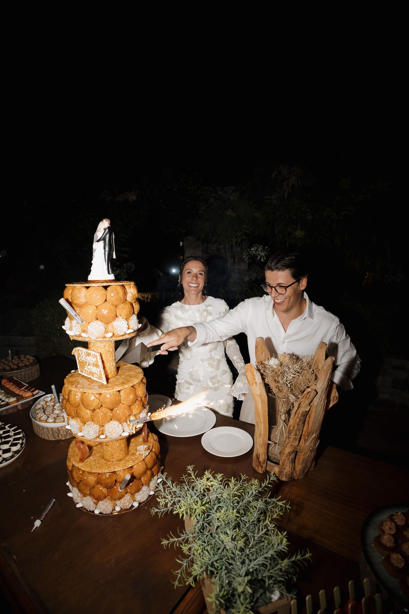 Bride and groom cutting a tiered croquembouche wedding cake outdoors at night.