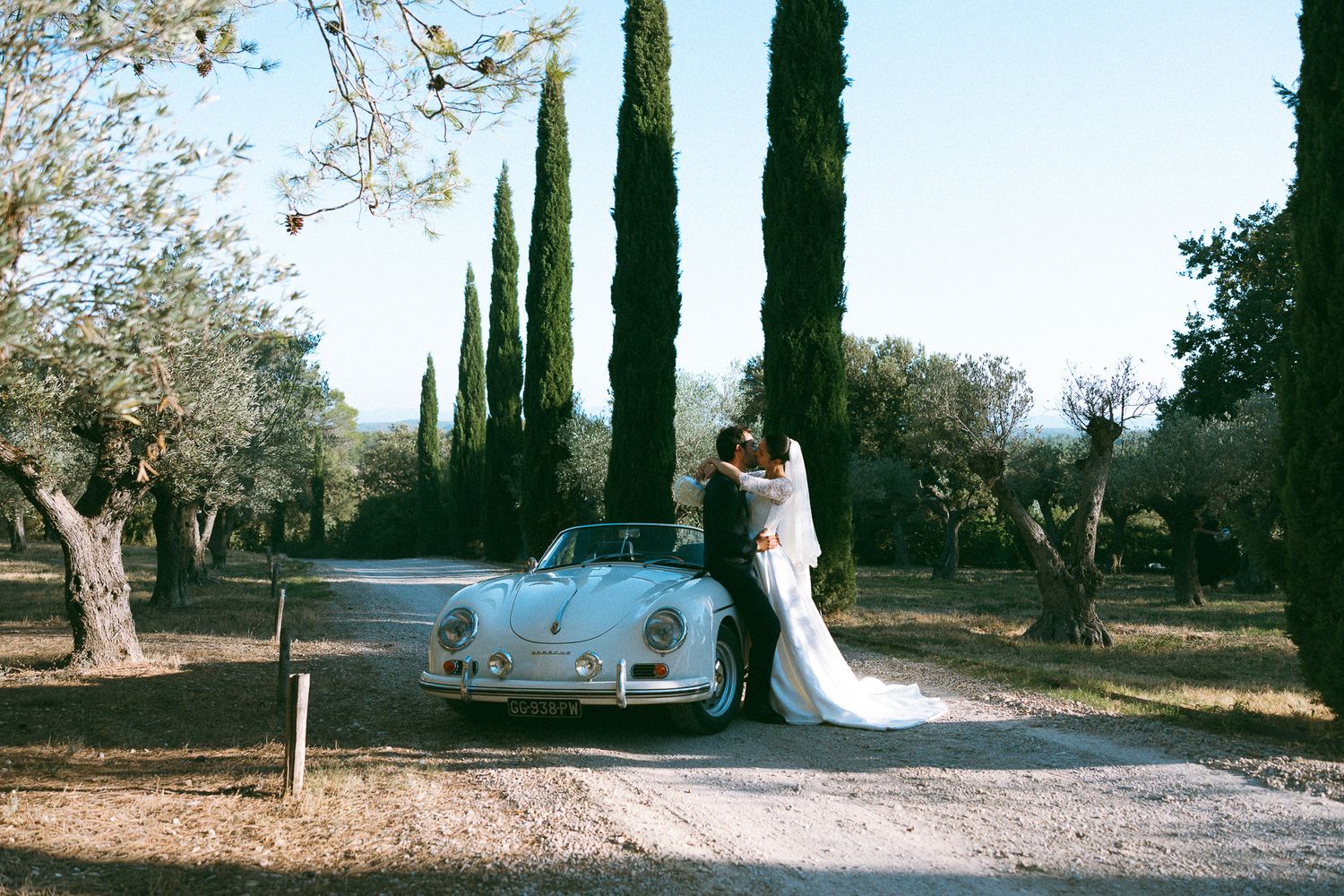 Bride and groom kissing beside a vintage white Porsche convertible surrounded by cypress trees and olive groves.