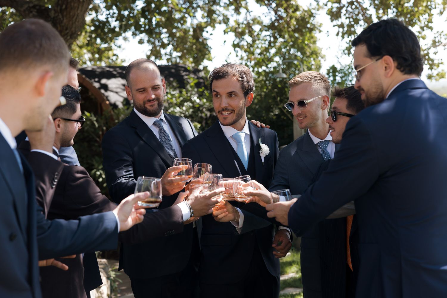 Groomsmen in suits toasting with wine glasses outdoors at a wedding celebration on a sunny day.