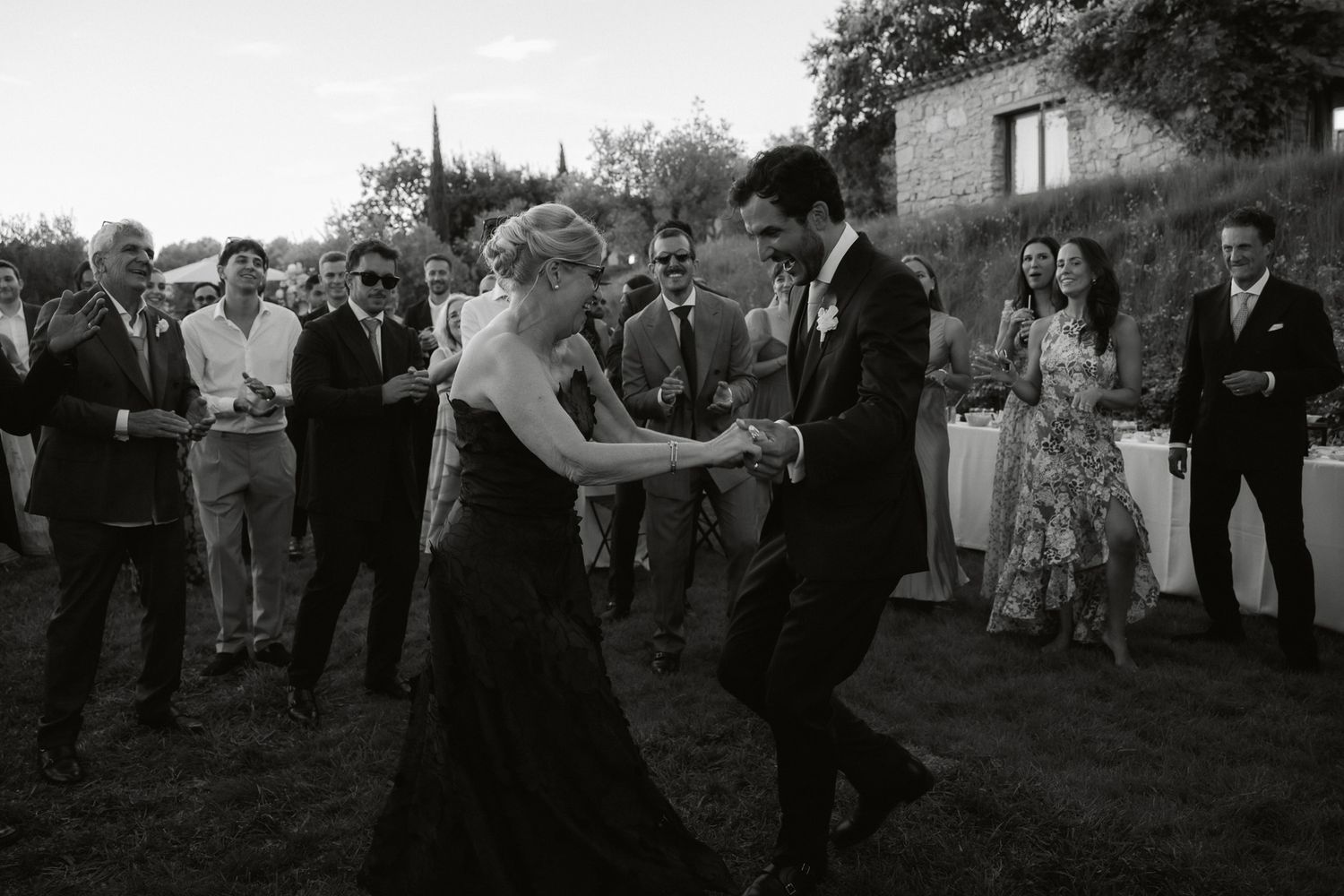 Black and white photo of bride and groom dancing joyfully outdoors at wedding reception with guests watching.