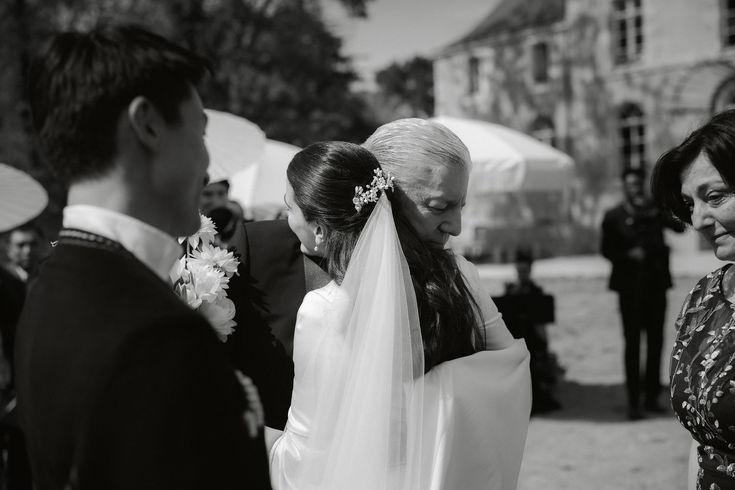 Black and white wedding photo of bride receiving a kiss from father during outdoor ceremony with guests watching.