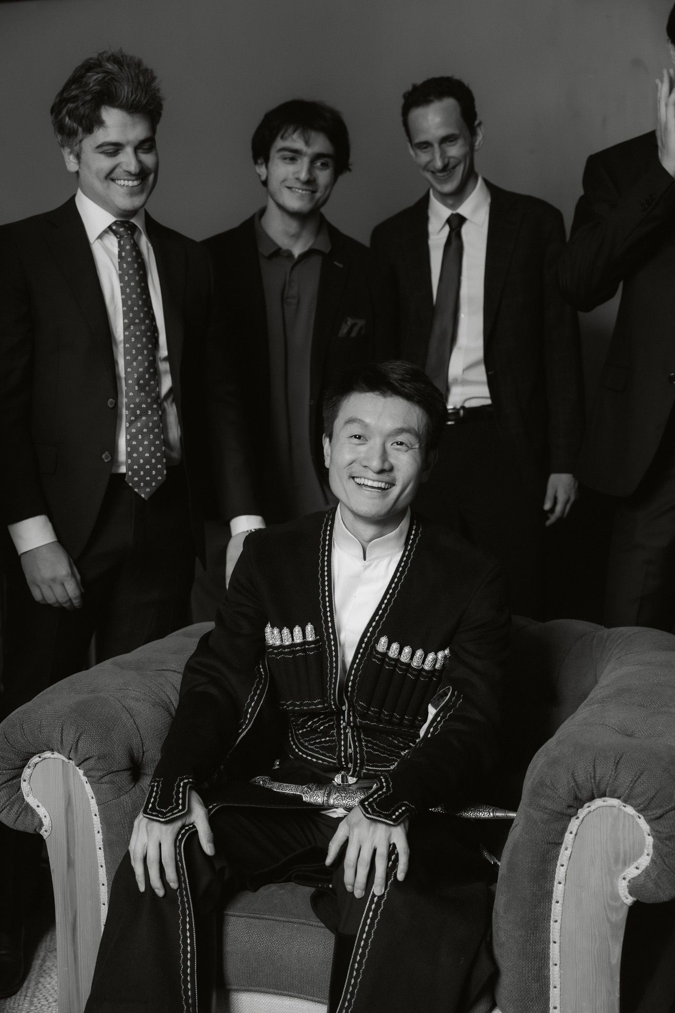 Black and white photo of four men in suits posing with one seated man in decorative traditional jacket.