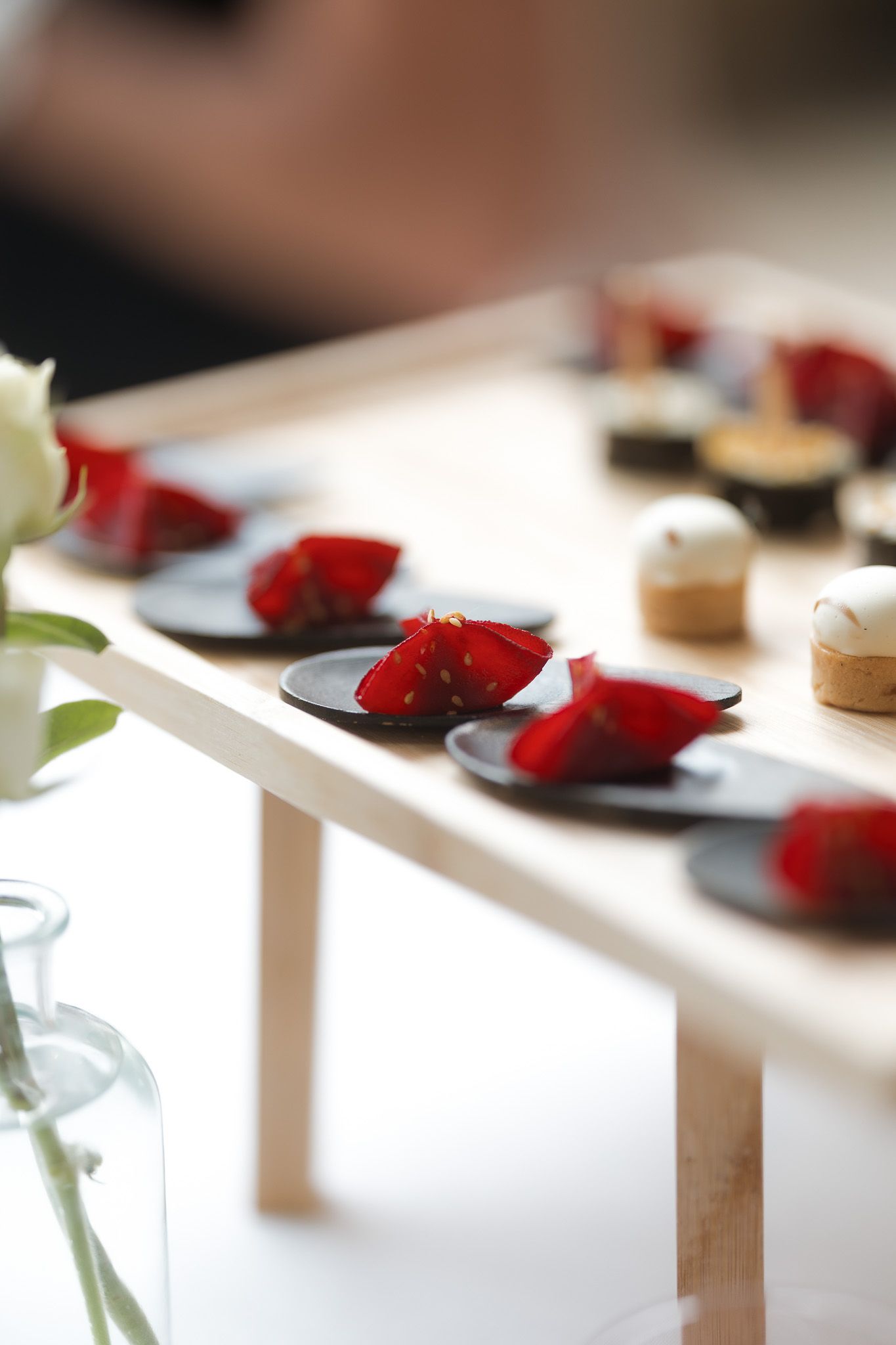Elegant wooden display stand with red flower petal desserts on black plates alongside pastries at a catered event.