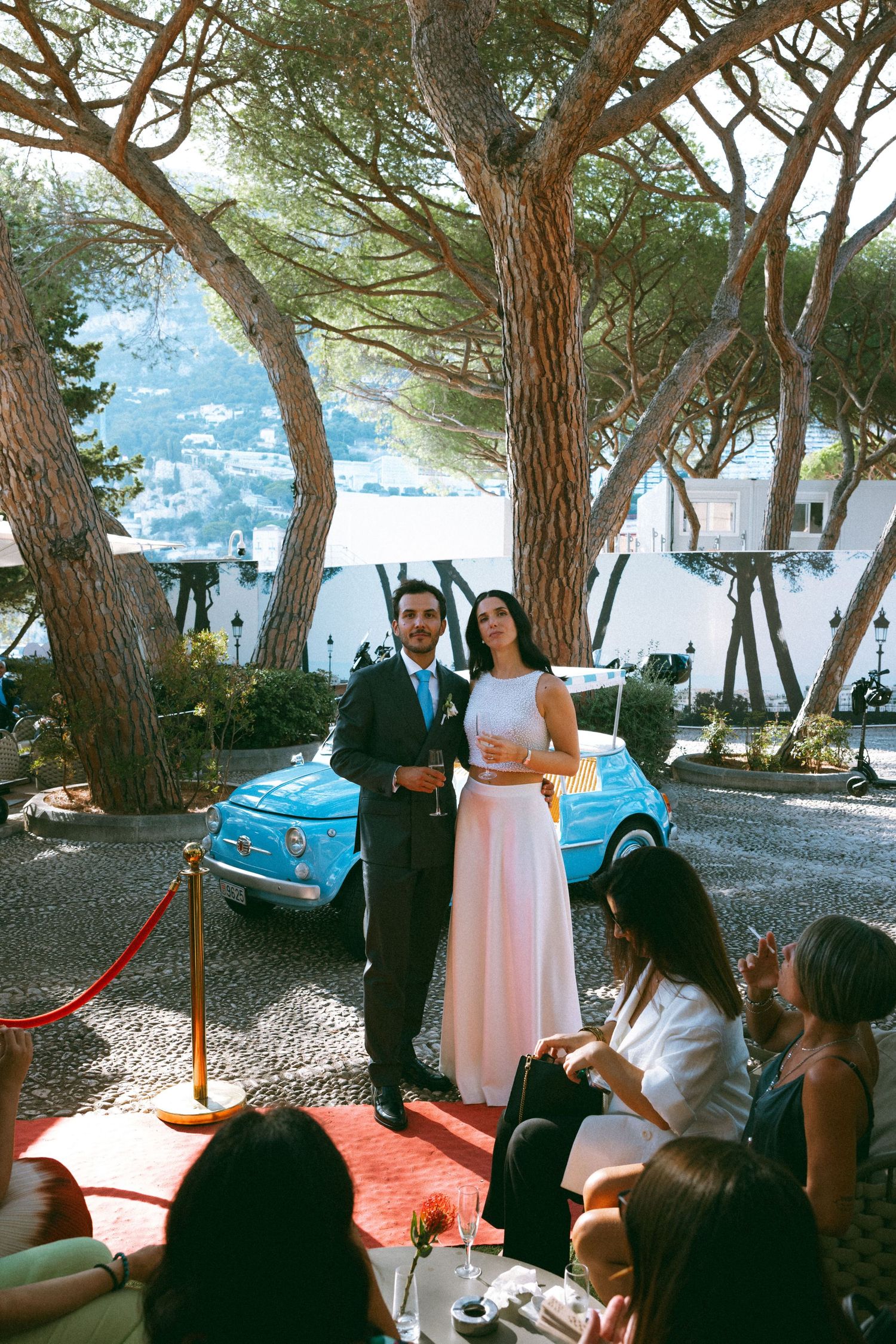 Couple in formal attire poses on red carpet beside vintage blue Fiat 500 under pine trees at Italian coastal venue.
