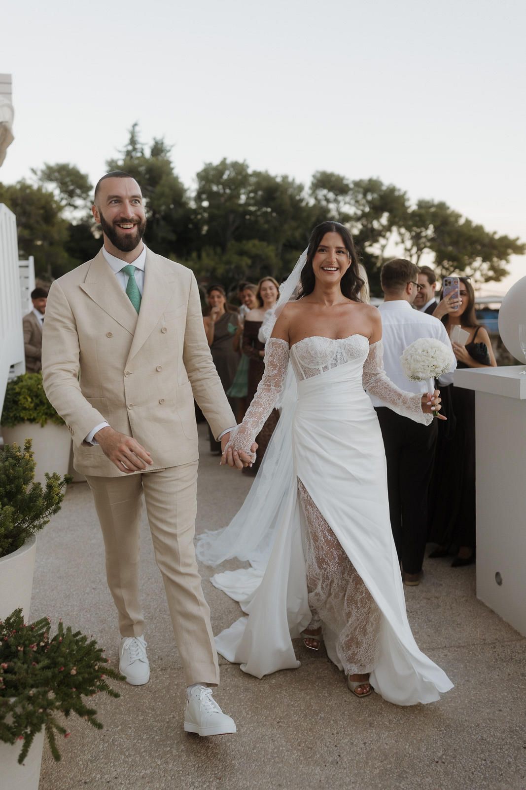A couple walks hand in hand at their outdoor wedding, with the groom in a beige suit and bride in a lace white gown.