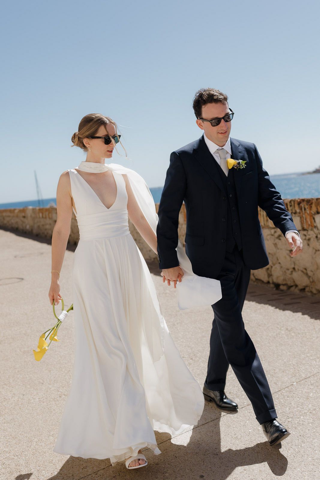 A couple in wedding attire walks hand in hand on a sunny beach path with a stone wall visible in the background.